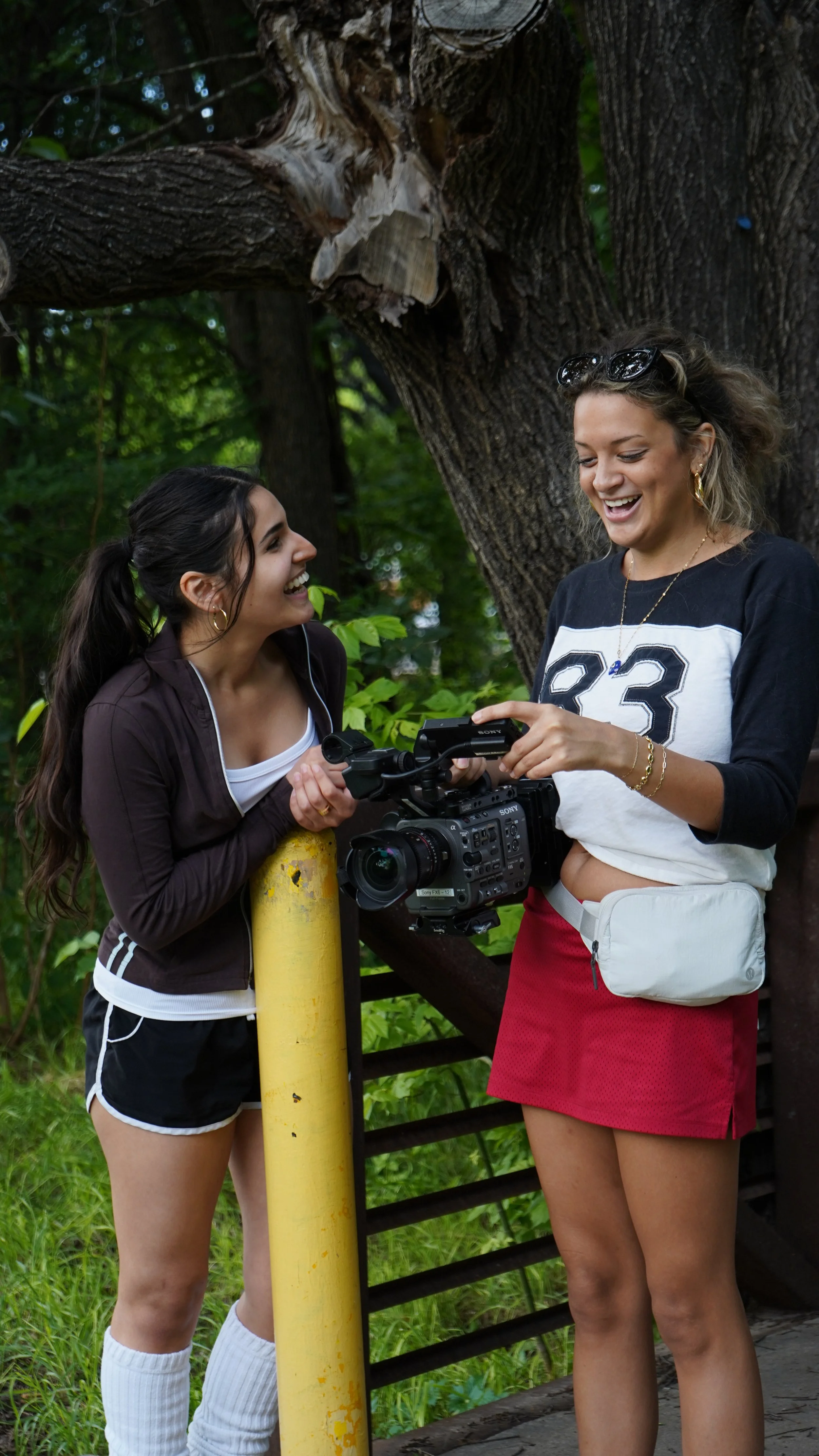 Two women share a laugh while filming outdoors with a professional video camera, standing near a large tree and greenery.