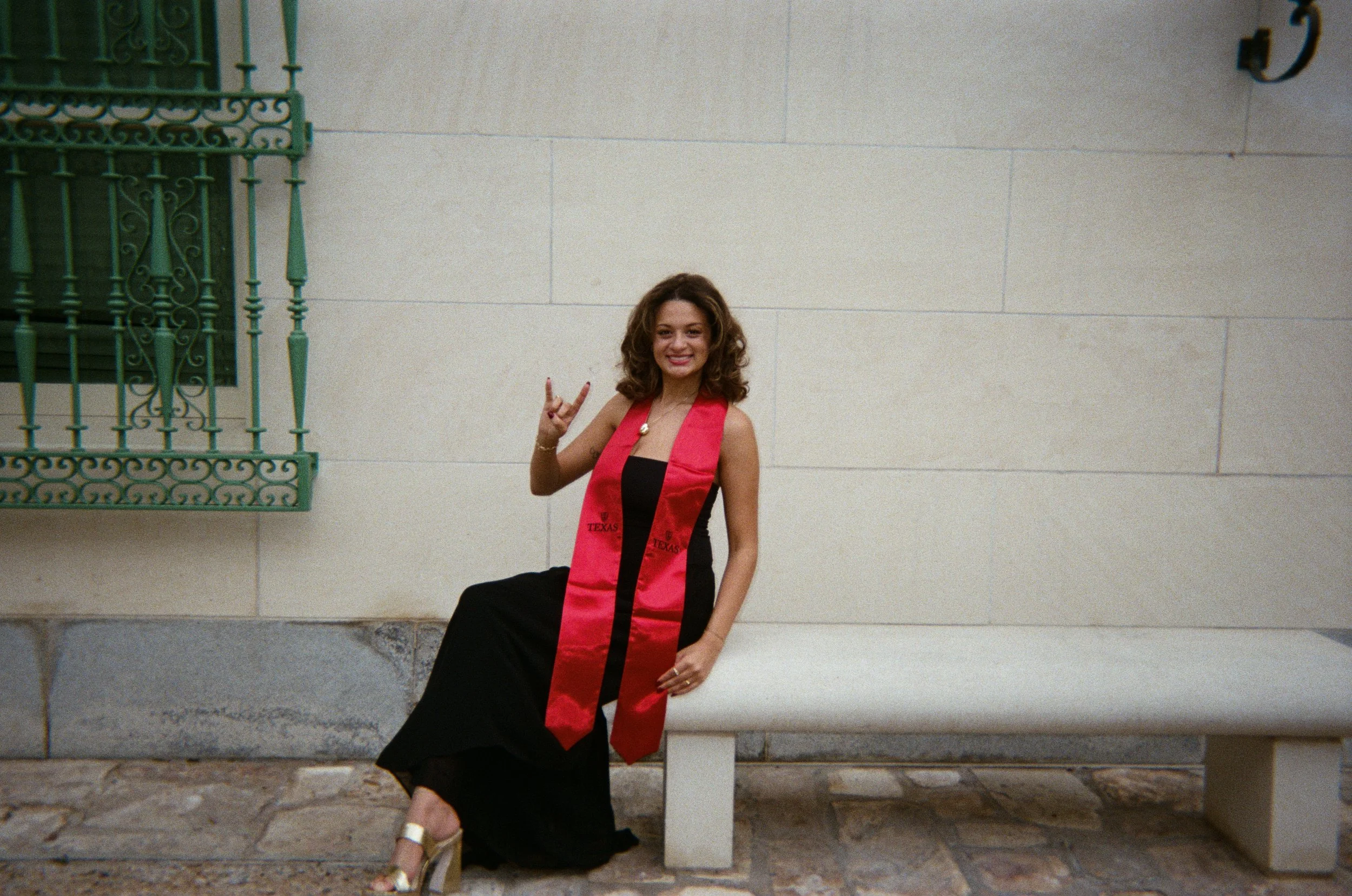 A young woman with curly hair sitting on a bench against a beige stone wall, wearing a graduation dress with a red stole labeled 'Texas' and smiling while making a peace sign.