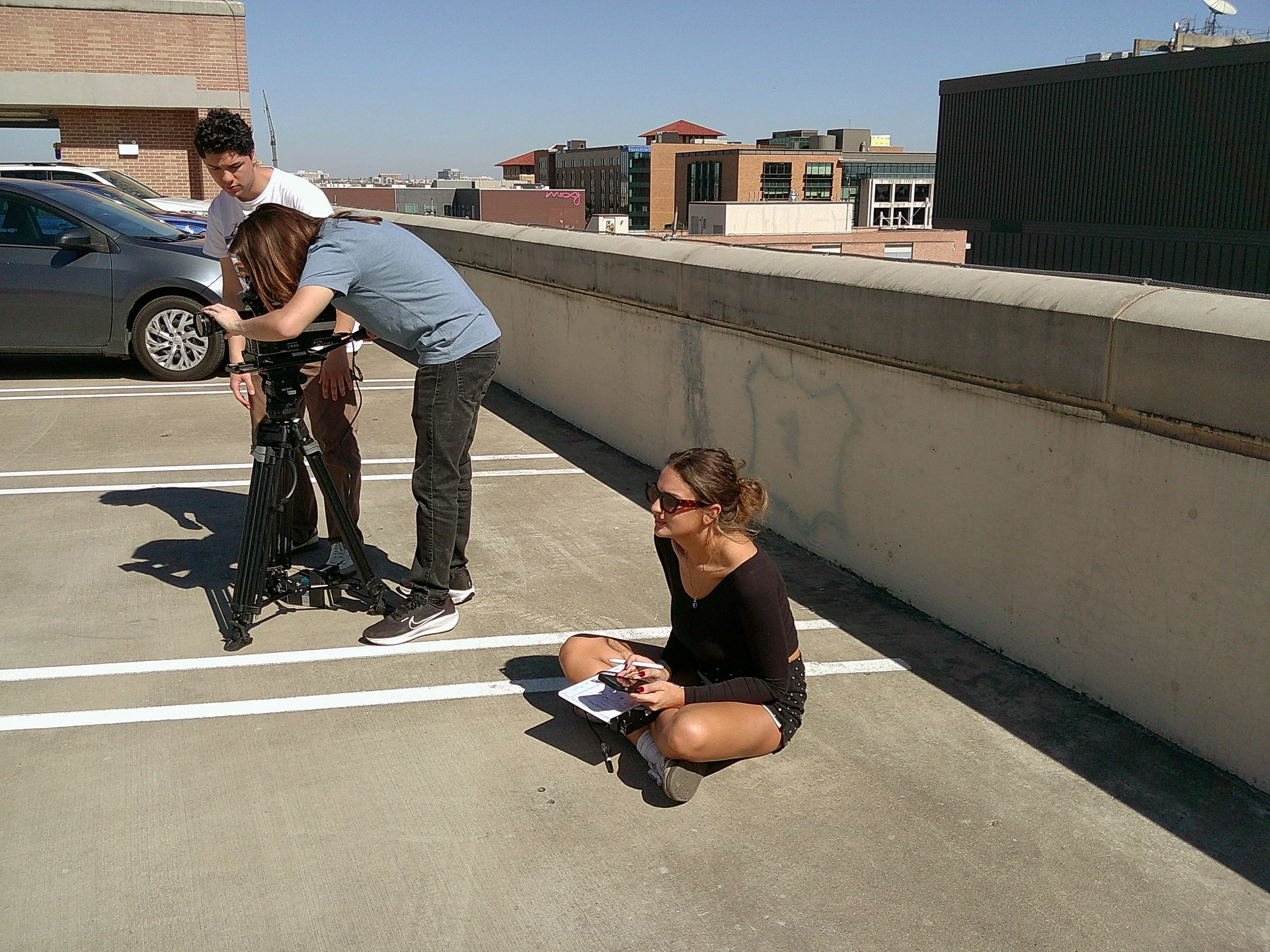 Three young women preparing filming equipment on the rooftop of a parking garage in an urban area.