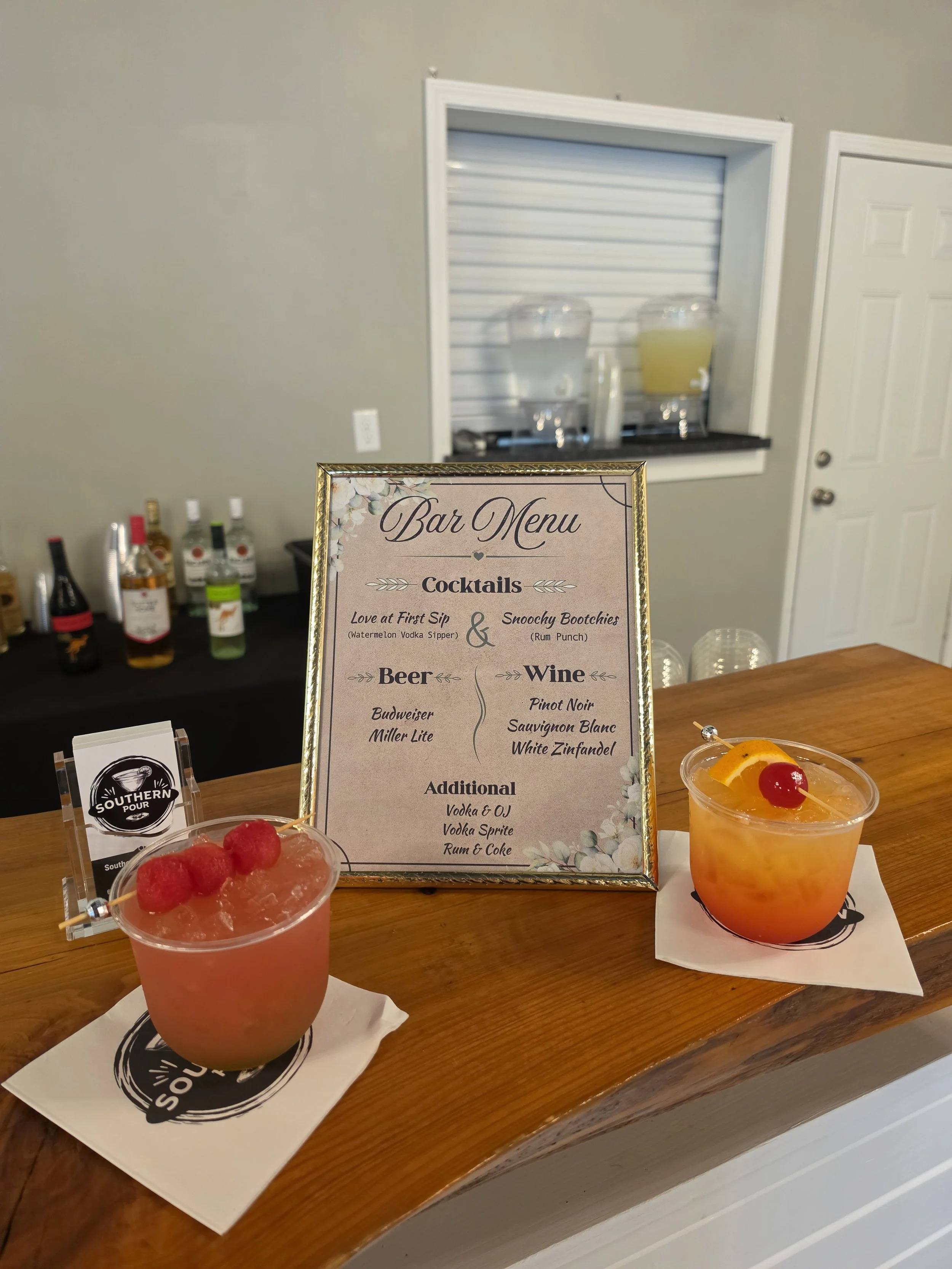 Two colorful cocktails with fruit garnishes on a wooden bar, with a cocktail menu sign and alcohol bottles in the background.