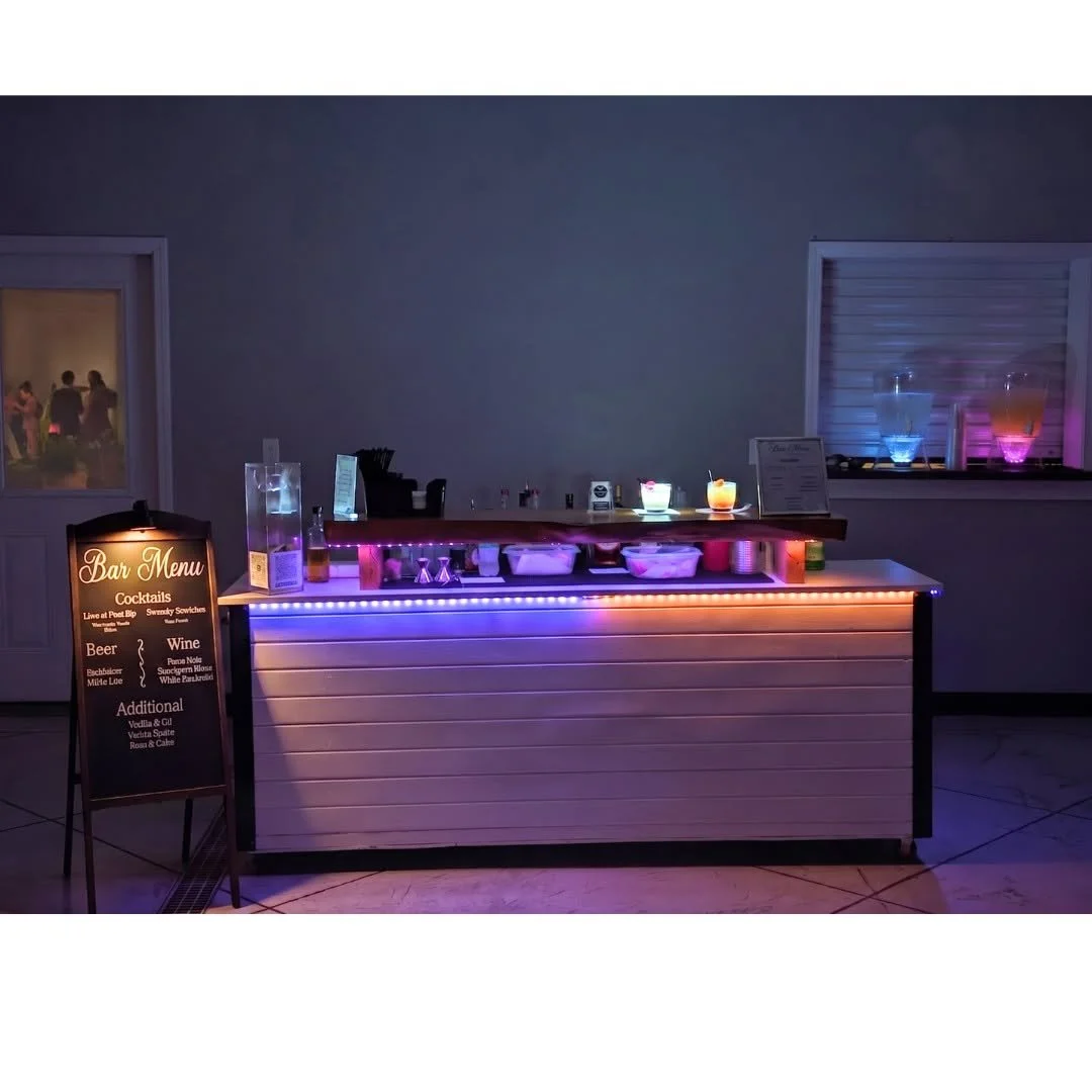 A lit-up bar counter with drinks and a chalkboard menu on the left listing cocktails, beer, wine, and additional drinks. Behind the counter are bottles and glasses, with a window showing people dancing in the background.