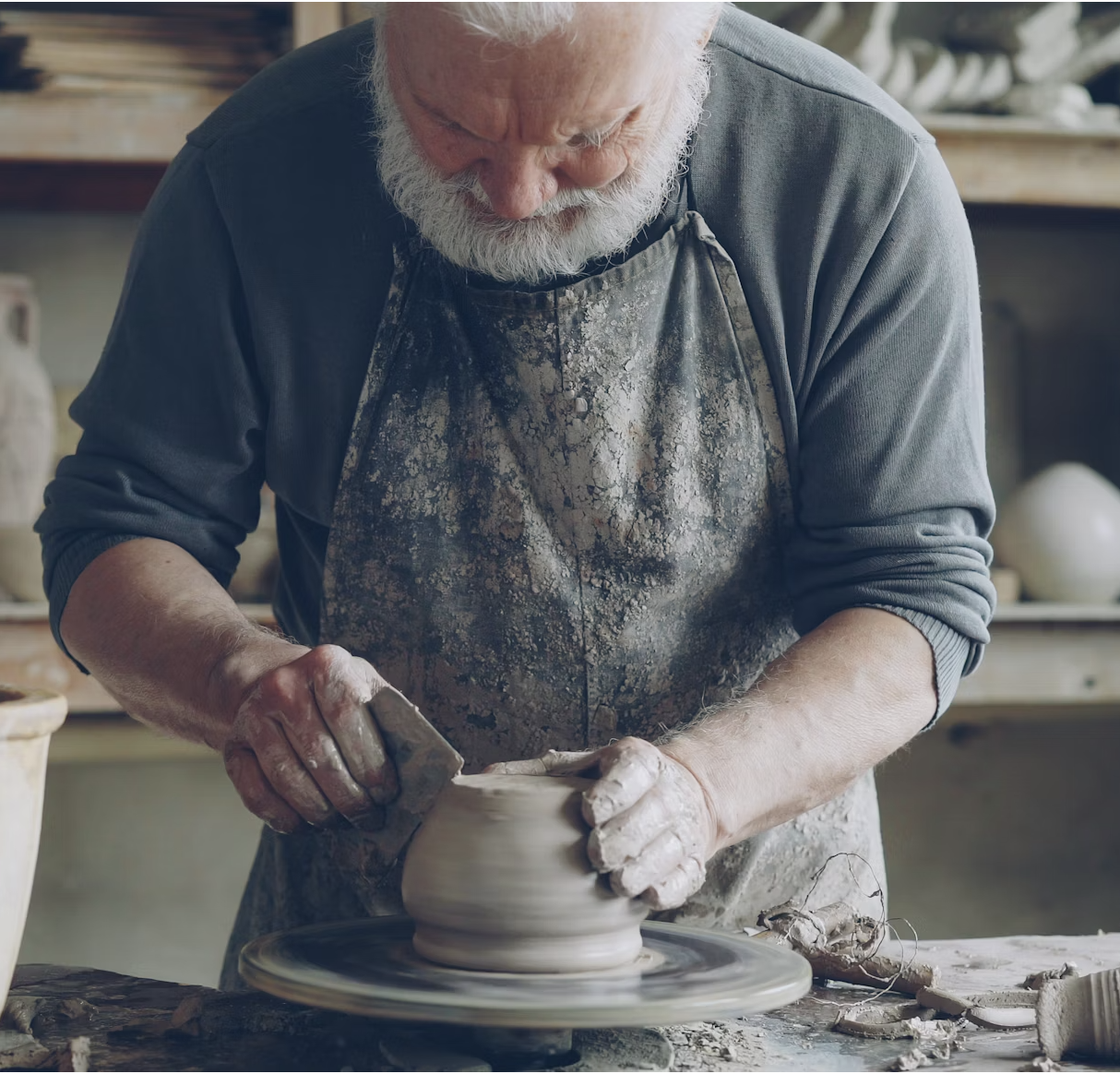 An elderly man with a white beard shaping a clay bowl on a pottery wheel in a pottery studio.