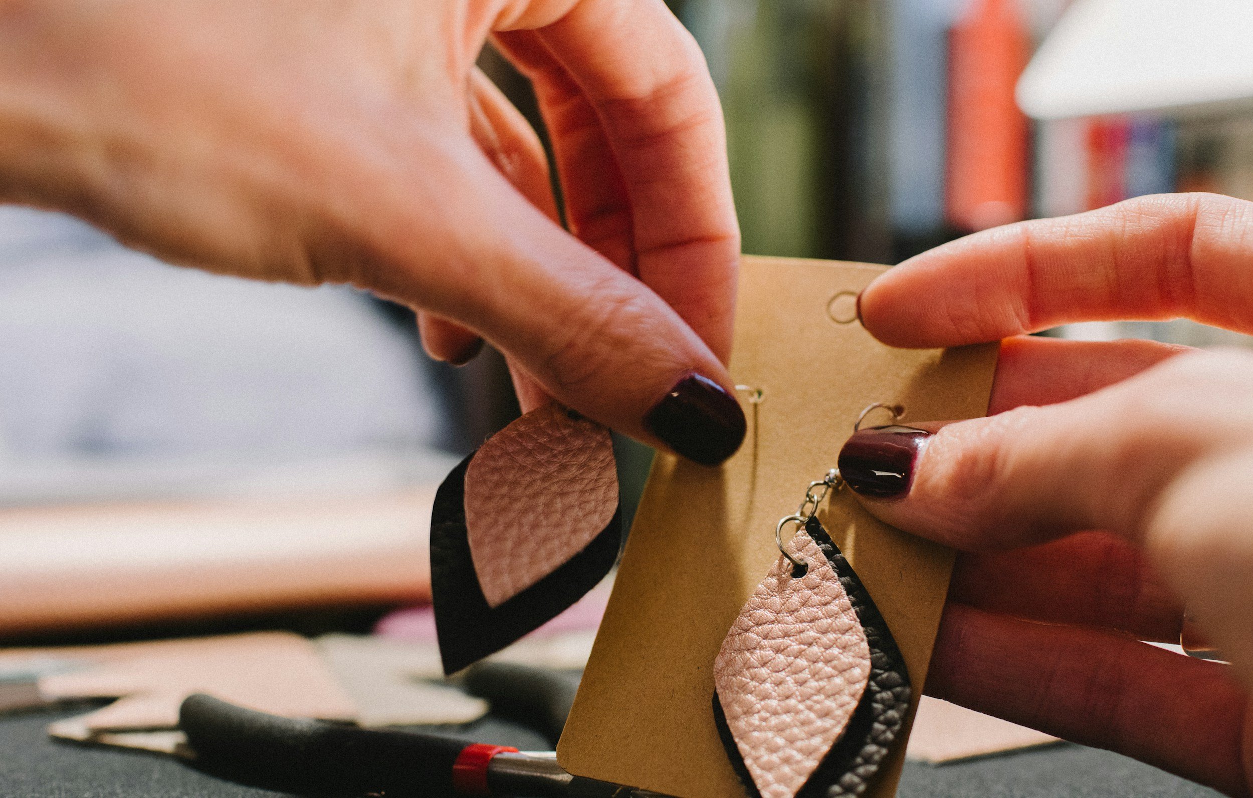 A person is holding a jewelry display card with handmade leather earrings, showing dark and light pink leather teardrops with black edges. The background includes tools and additional jewelry pieces.