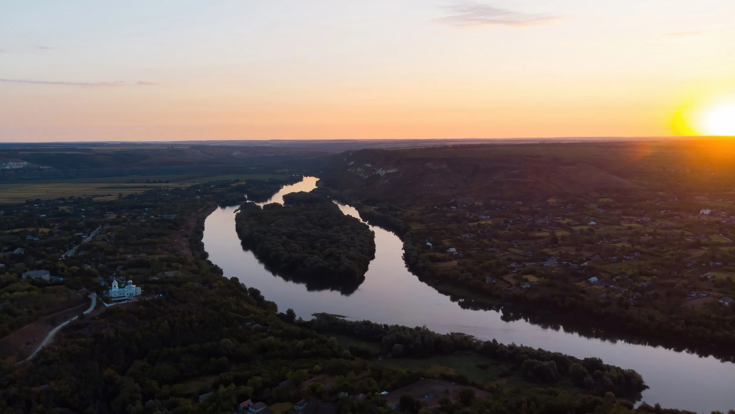 Aerial view of a winding river flowing through a rural landscape at sunset, with a church and small houses along the riverbanks.