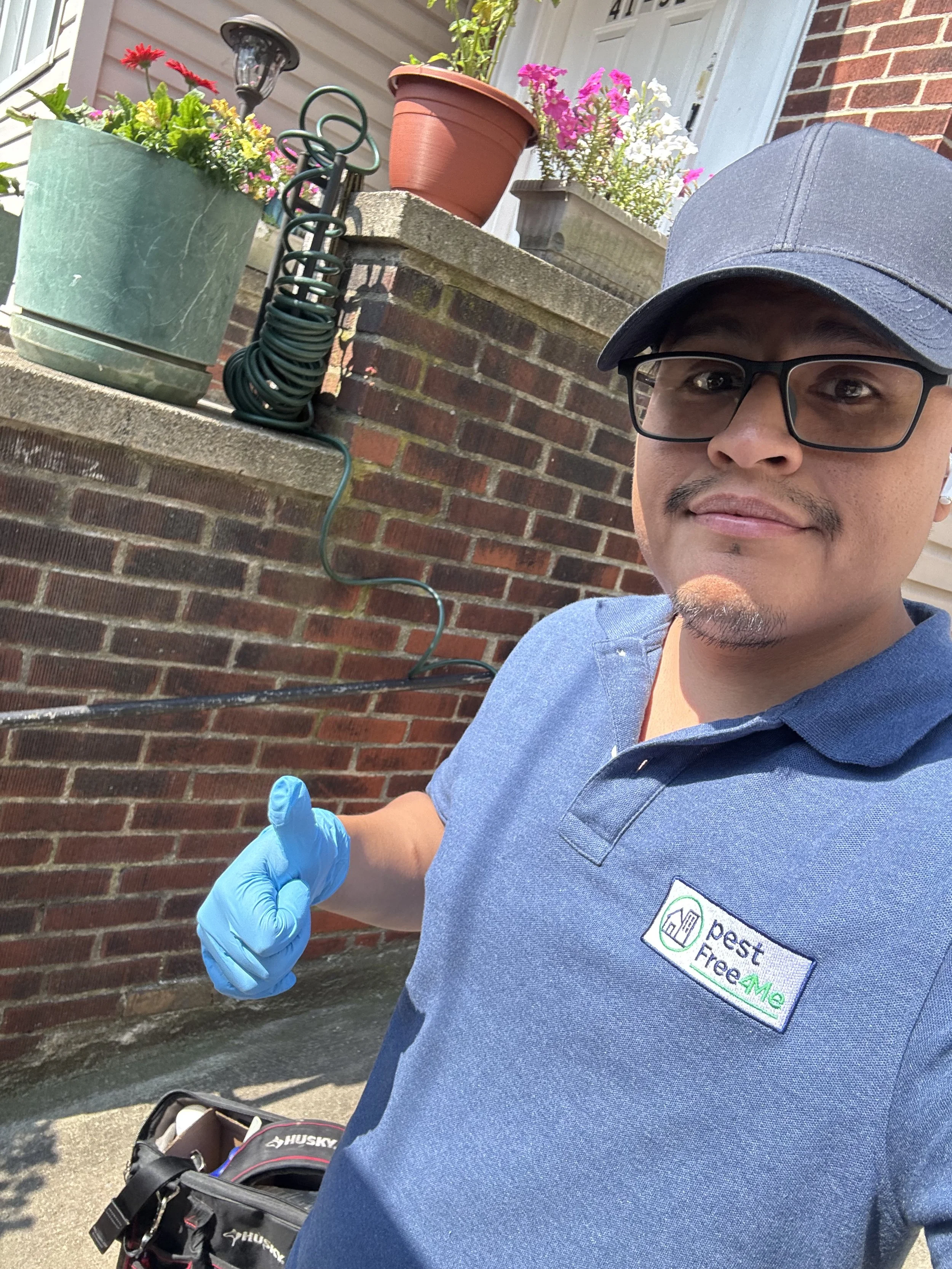A man taking a selfie outdoors in front of a brick wall with potted plants and gardening tools. He is wearing a blue polo shirt with a logo that reads 'pest Free & Me' and a dark cap. He has glasses, a slight mustache, and is wearing blue gloves.