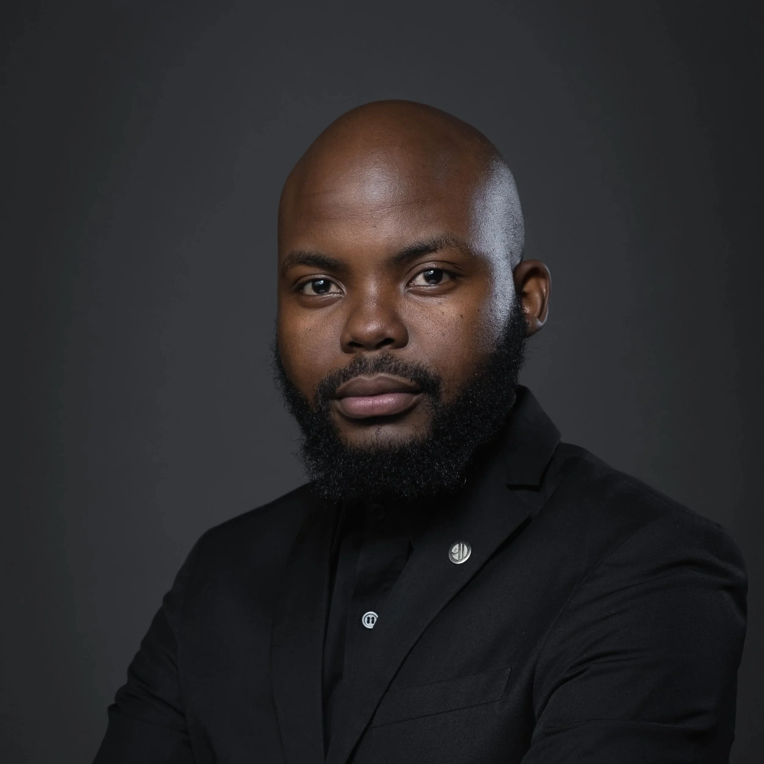 Portrait of a man with a beard, wearing a black suit with a shirt, standing against a dark background.