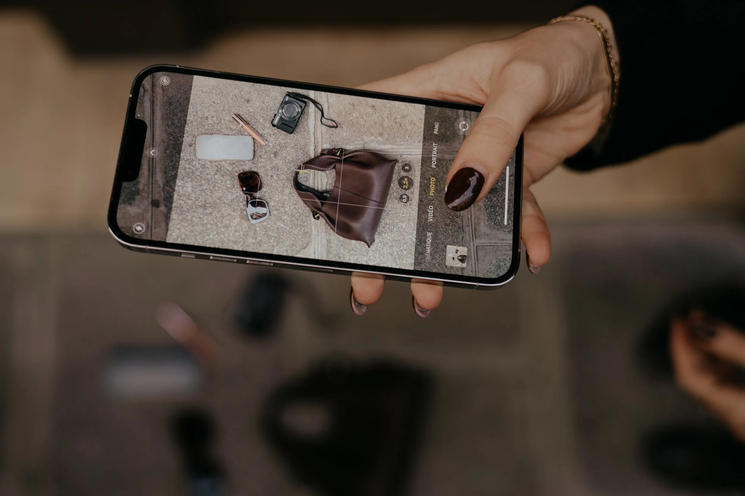 A person holding a smartphone takes a photo of a collection of accessories and personal items placed on a beige carpet, including sunglasses, a handbag, a camera, and a lipstick.