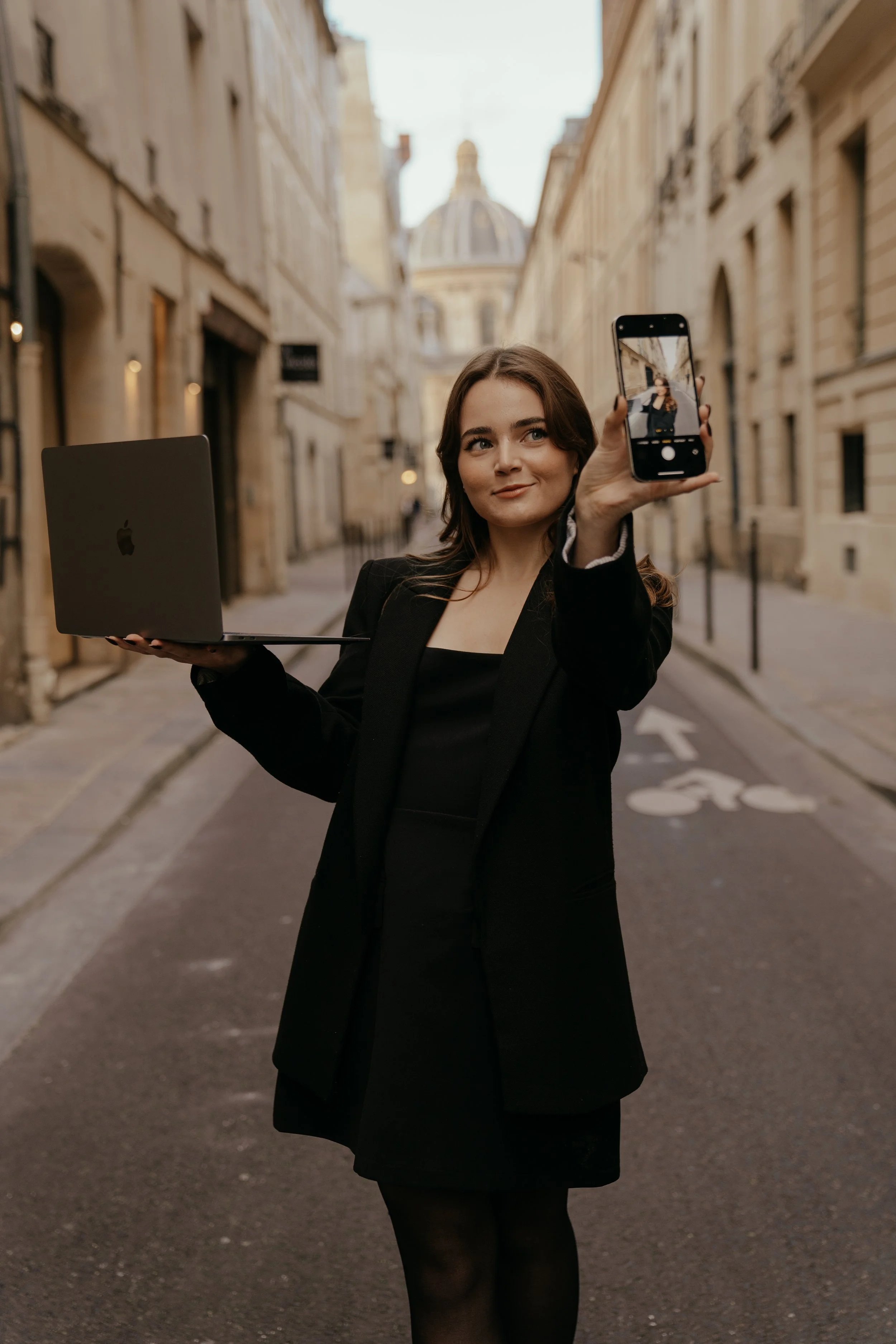 A woman standing on a street in Paris, holding a laptop in one hand and taking a selfie with her phone in the other, with historic buildings and a domed structure in the background.