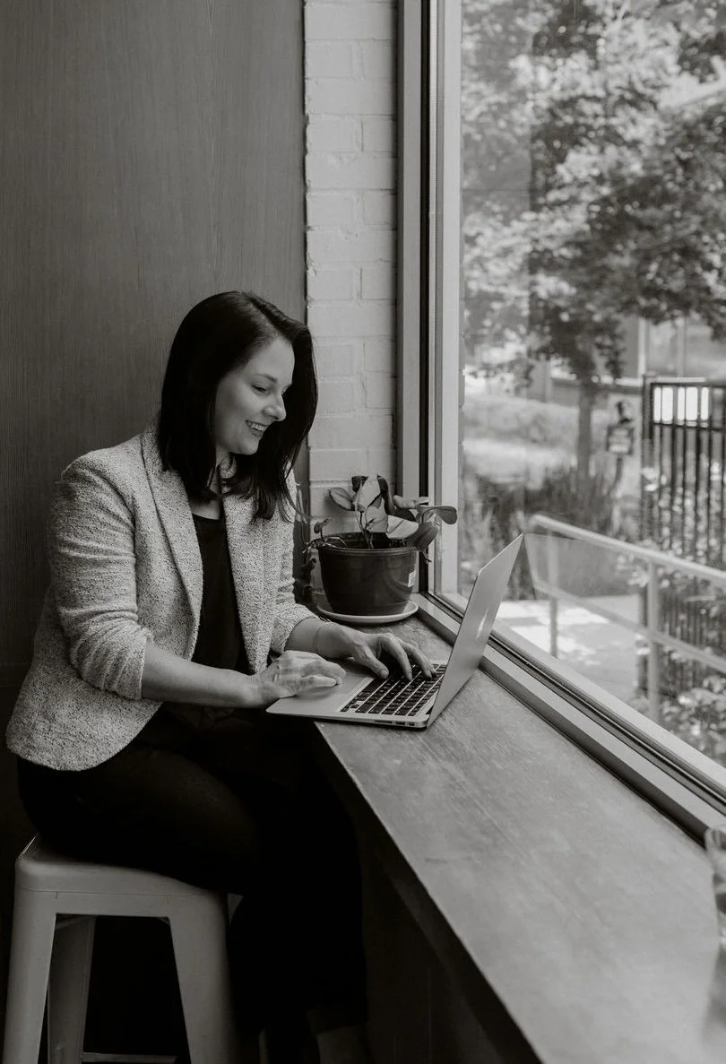 A woman with dark hair sitting by a large window, using a laptop on a wooden windowsill, with a potted plant beside her, smiling as she looks at the screen.