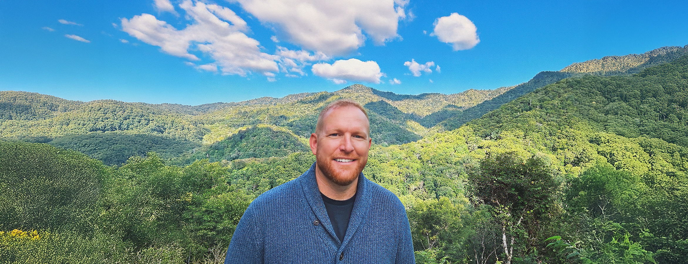 A smiling man with a beard wearing a blue sweater standing outdoors in front of a lush green mountain landscape with a blue sky and white clouds.