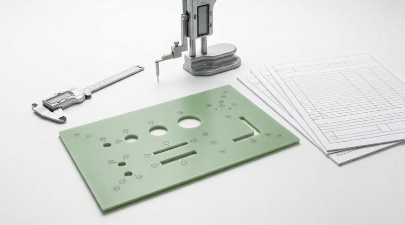 A green circuit board with multiple holes and markings, a pair of calipers, a robotic arm, and stacks of paper with graphs on a white surface.