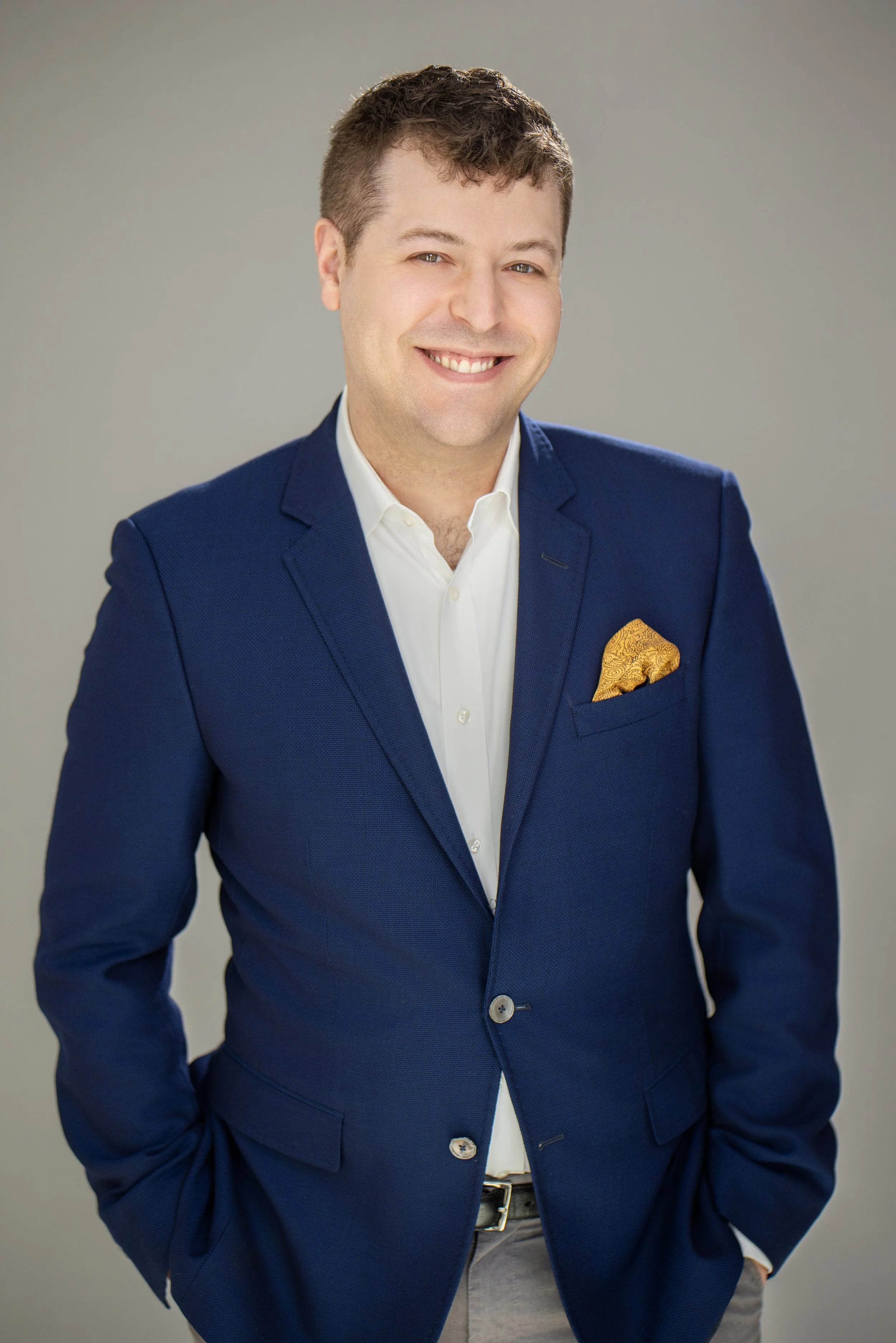David Davis, a smiling music educator in a navy blue blazer, white shirt, with a gold pocket square, standing against a plain background.