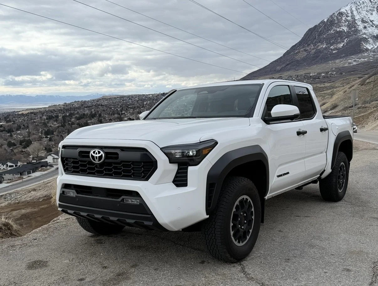 A white Toyota Tacoma pickup truck parked on a mountain road with mountainous landscape and cloudy sky in the background.