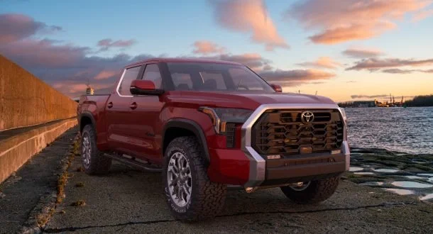 Red Toyota pickup truck parked by the water during sunset with a cloudy sky.