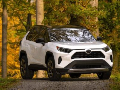 A white Toyota SUV parked on a gravel path in a forest with trees showing fall foliage in the background.