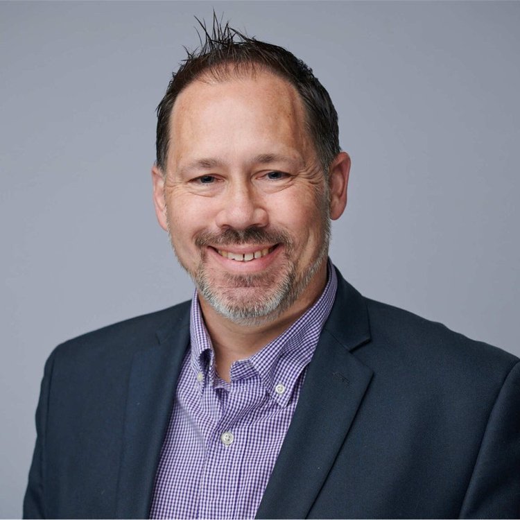 A man with short brown hair, a beard, and mustache wearing a dark blazer and a purple checkered shirt smiling at the camera against a gray background.