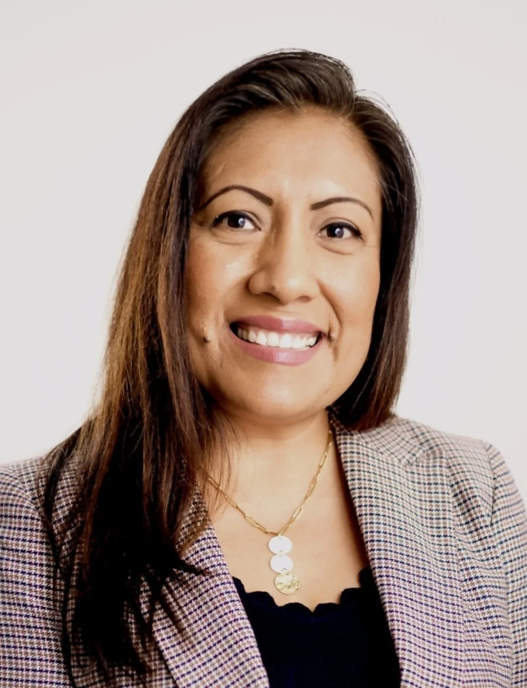 A woman with long brown hair smiling, wearing a beige checked blazer, black top, and gold necklace with coin pendants.