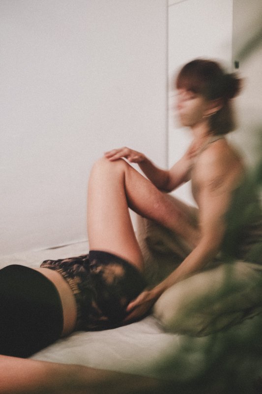 Zwei Menschen befinden sich in einer Massage-Session in einem Raum mit weißer Wand. Einer liegt auf dem Rücken, die andere Person sitzt neben ihm und berührt ihn.