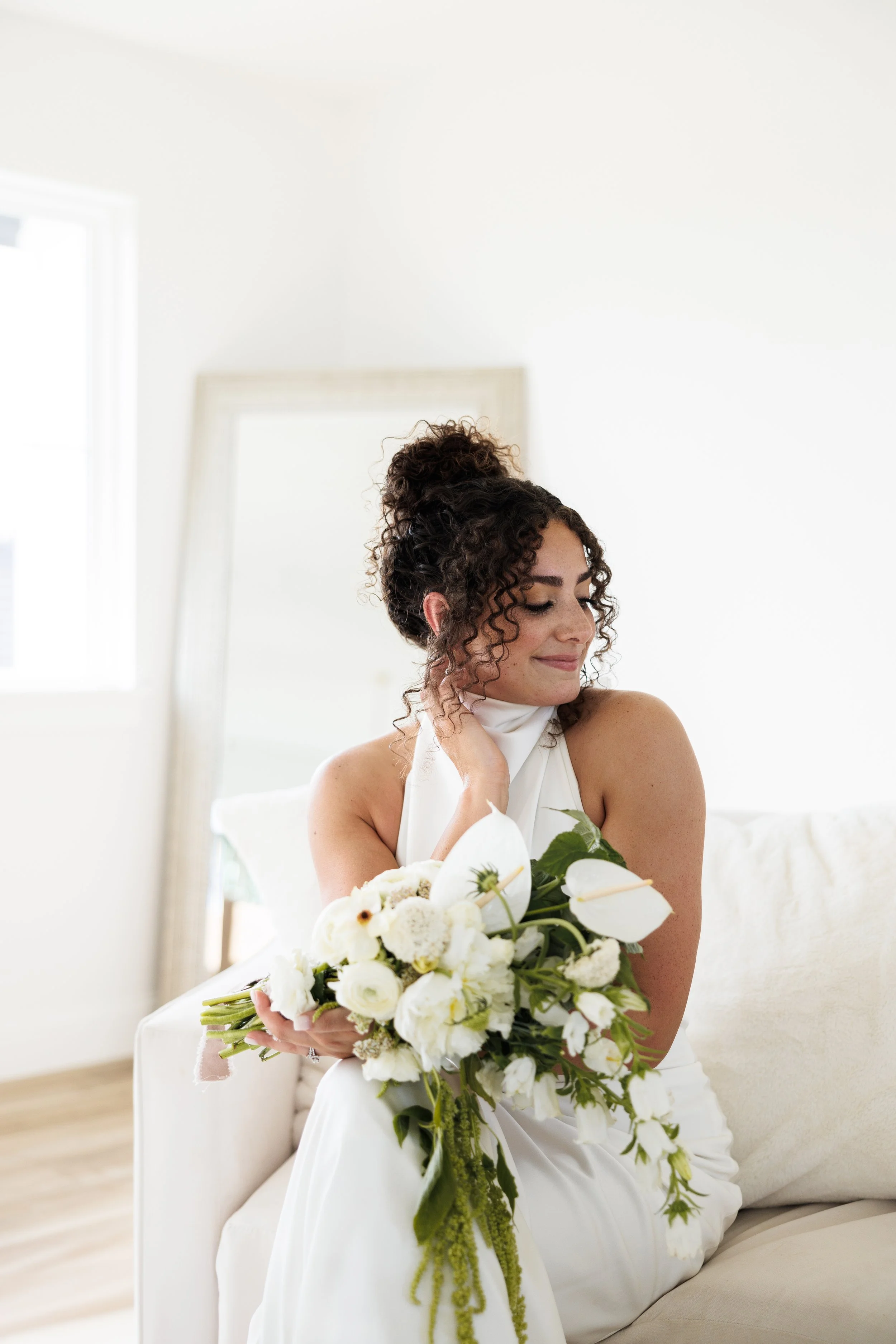 A woman in a white dress sitting on a couch, holding a bouquet of white flowers, with curly hair styled up, in a bright room.