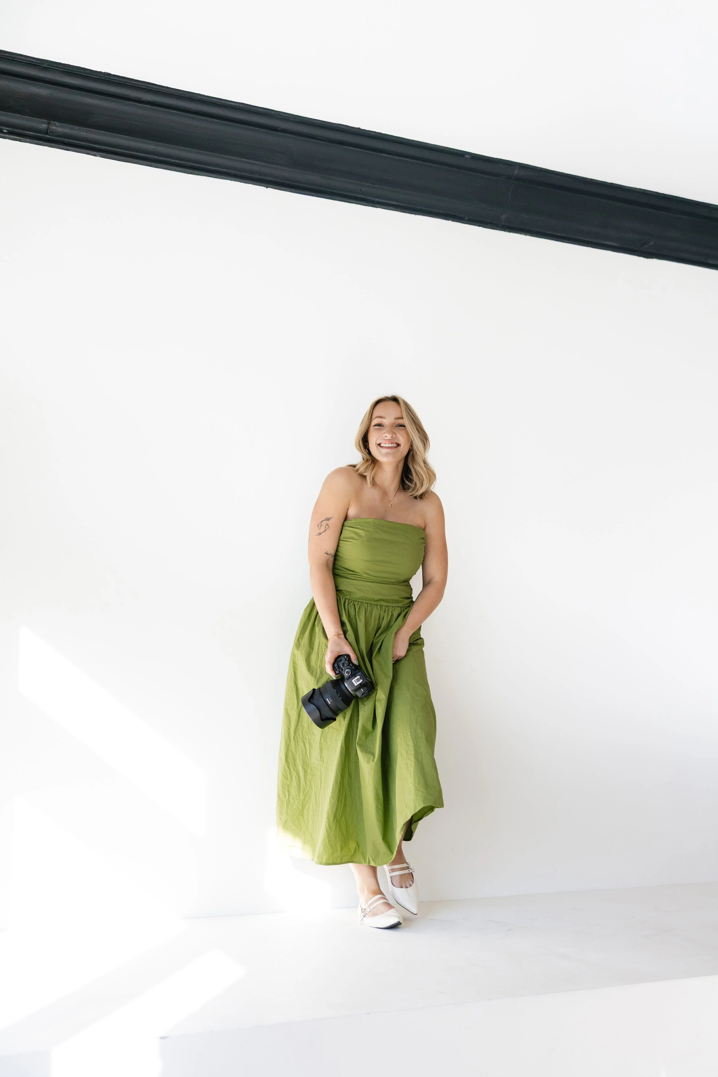 A woman in a green strapless dress holding a camera, standing against a white background in a photography studio.