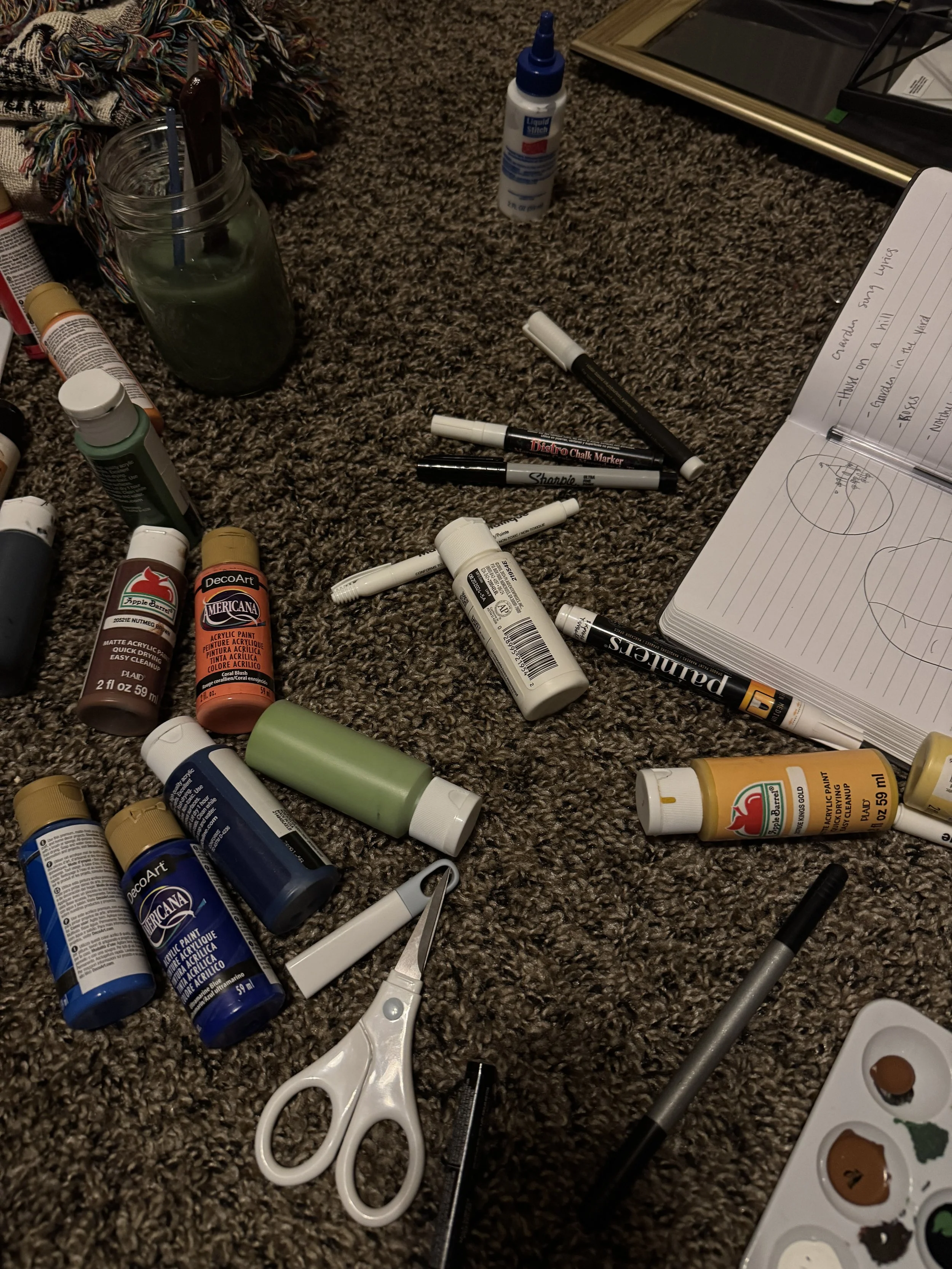 Assorted art supplies on a carpeted floor, including acrylic paints, markers, glue, scissors, a notebook, and a watercolor palette.