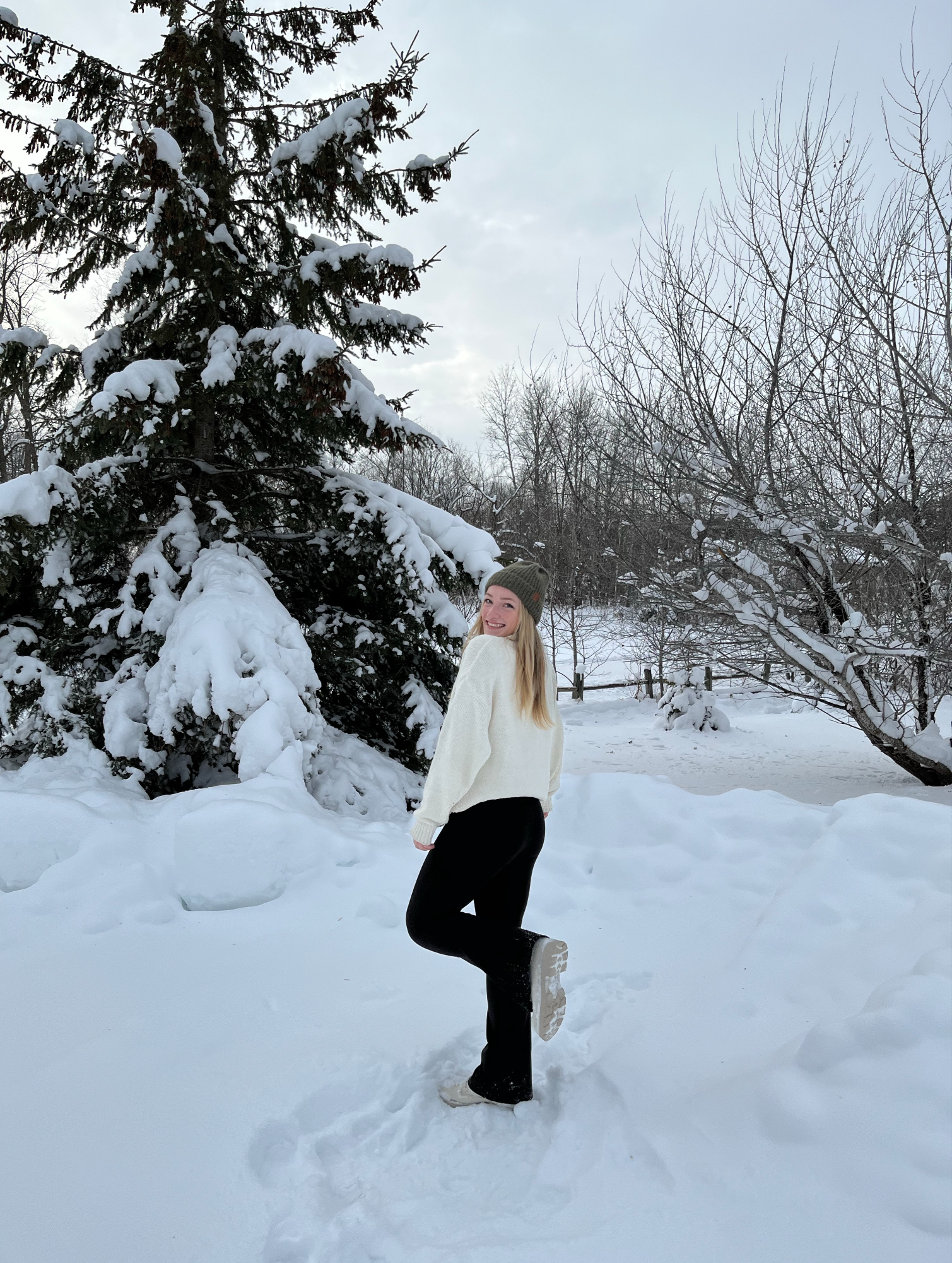 A smiling woman in a white sweater, black pants, white shoes, and a green knit hat standing on snow with one foot raised behind her, in a winter landscape with snow-covered trees and a cloudy sky.