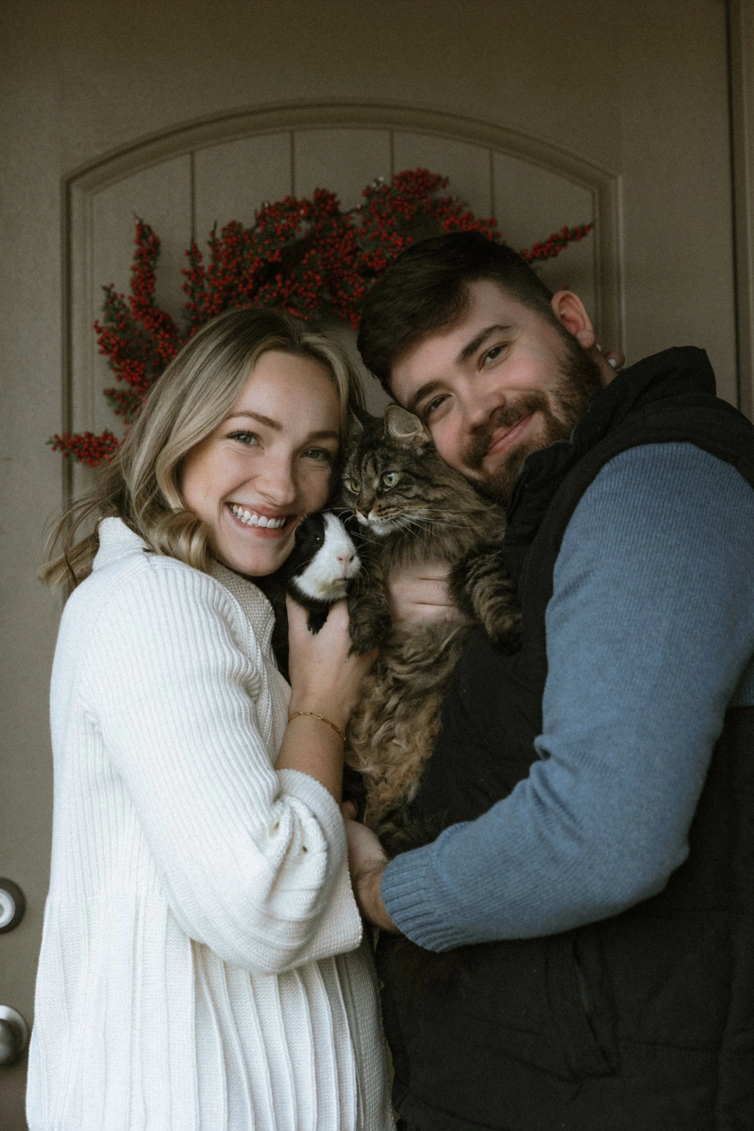 A smiling couple holds two cats in front of a holiday wreath on a door.