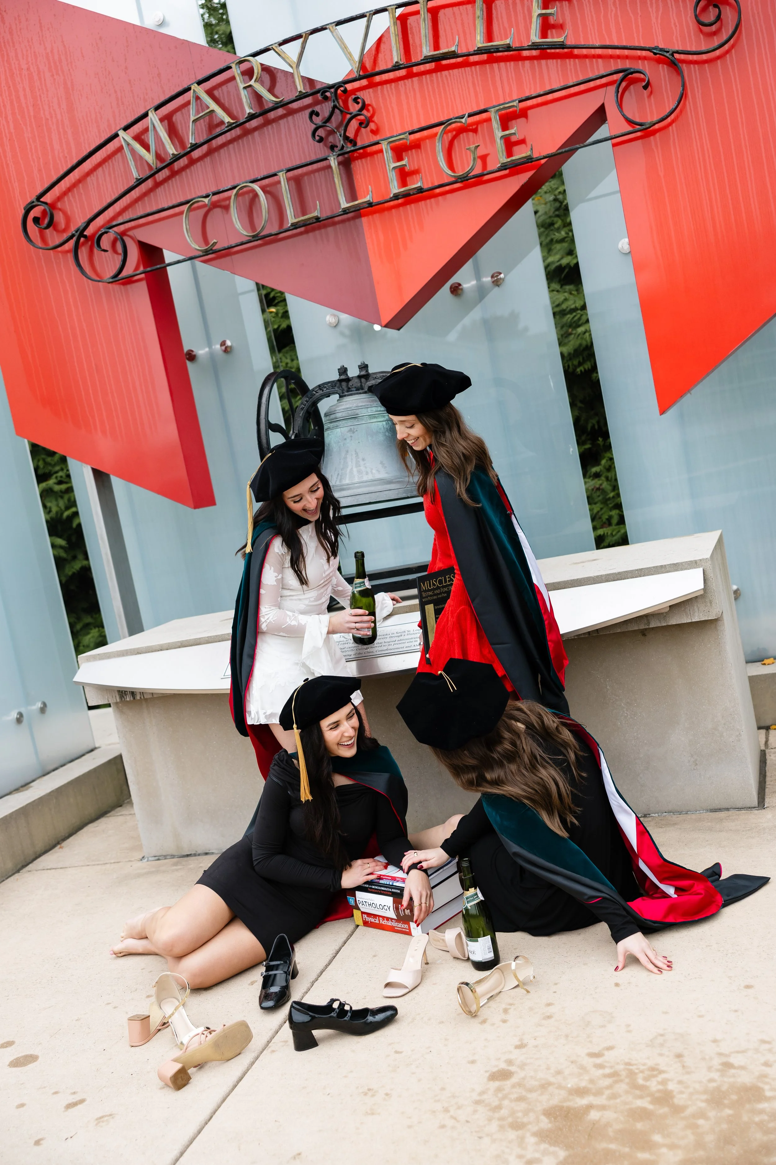 Graduates in caps and gowns celebrating on the ground outside a college building with a large bell and sign reading 'Marryville College'
