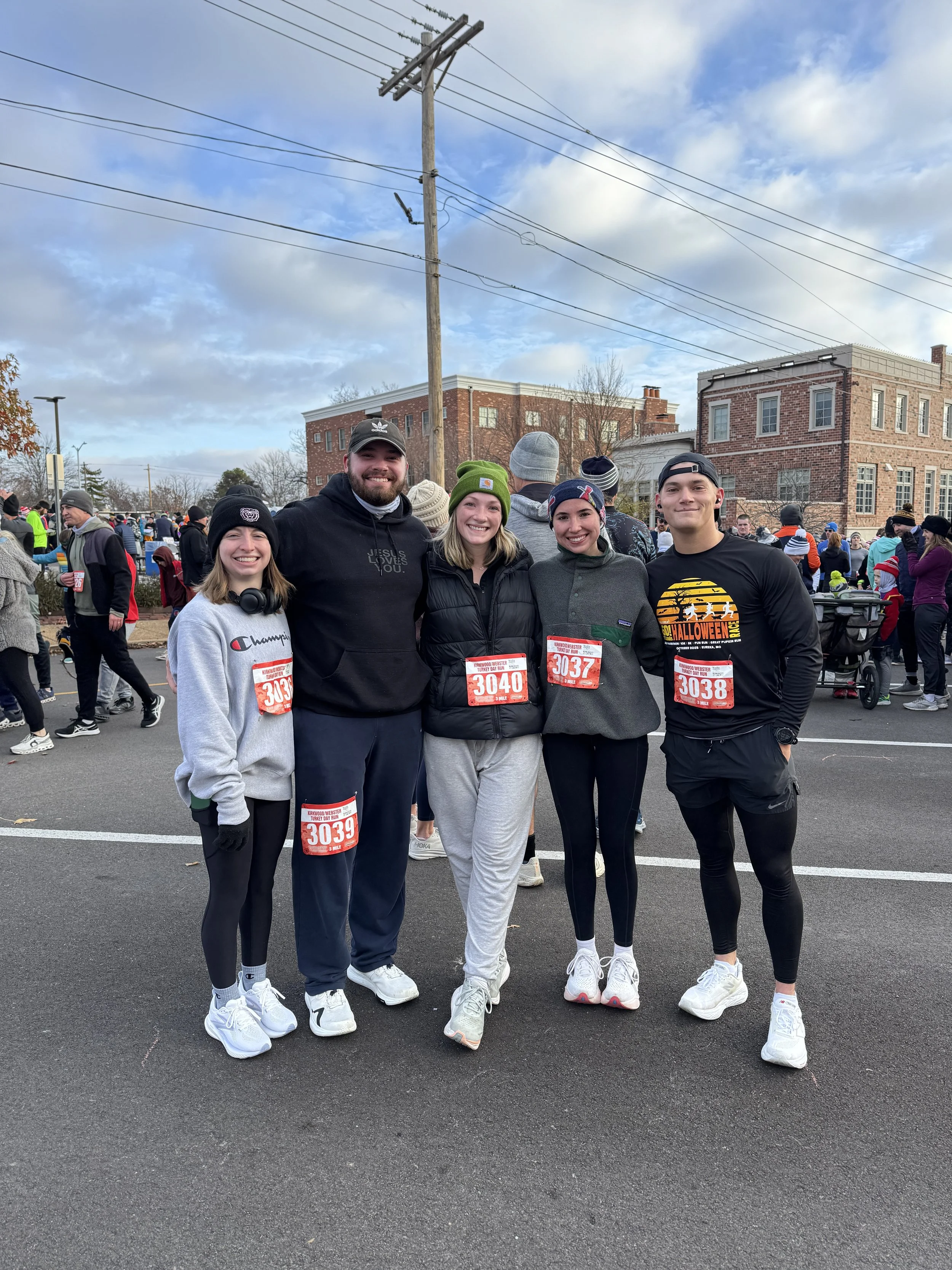 A group of five runners standing together, smiling at a race event, with other participants and officials in the background, in a parking lot and city street scene.