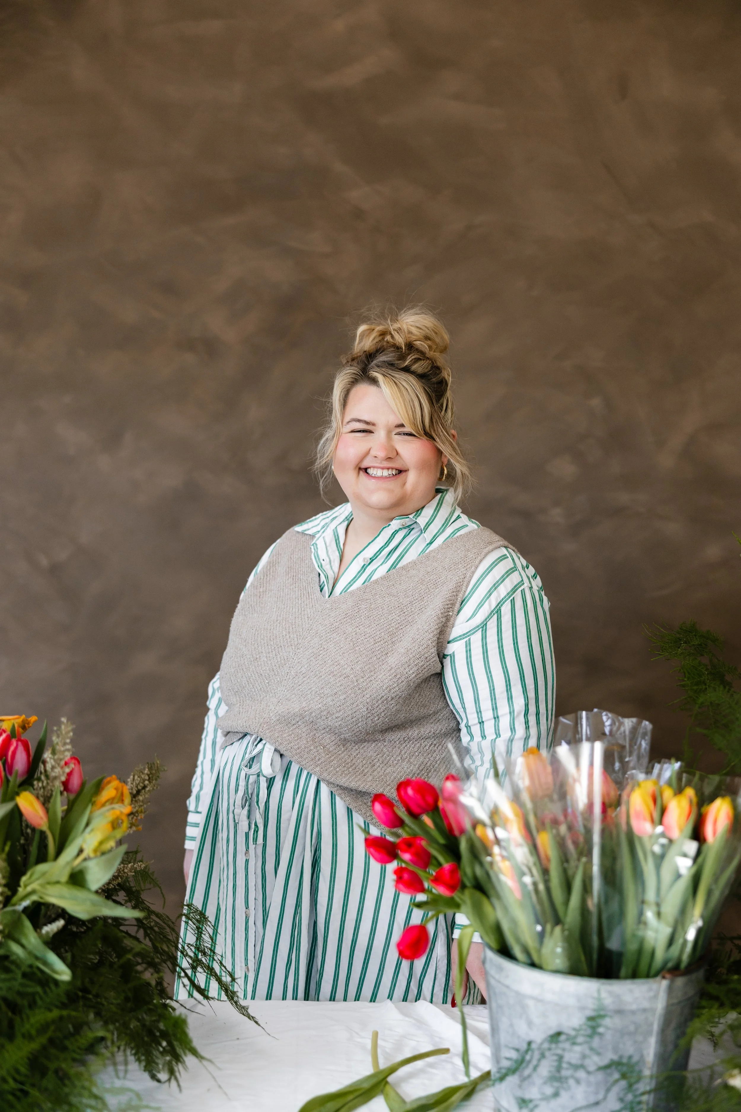 A smiling woman with a top bun hairstyle standing behind a table of colorful tulips and flowers in a floral shop or market. She is wearing a striped shirt and a beige vest.