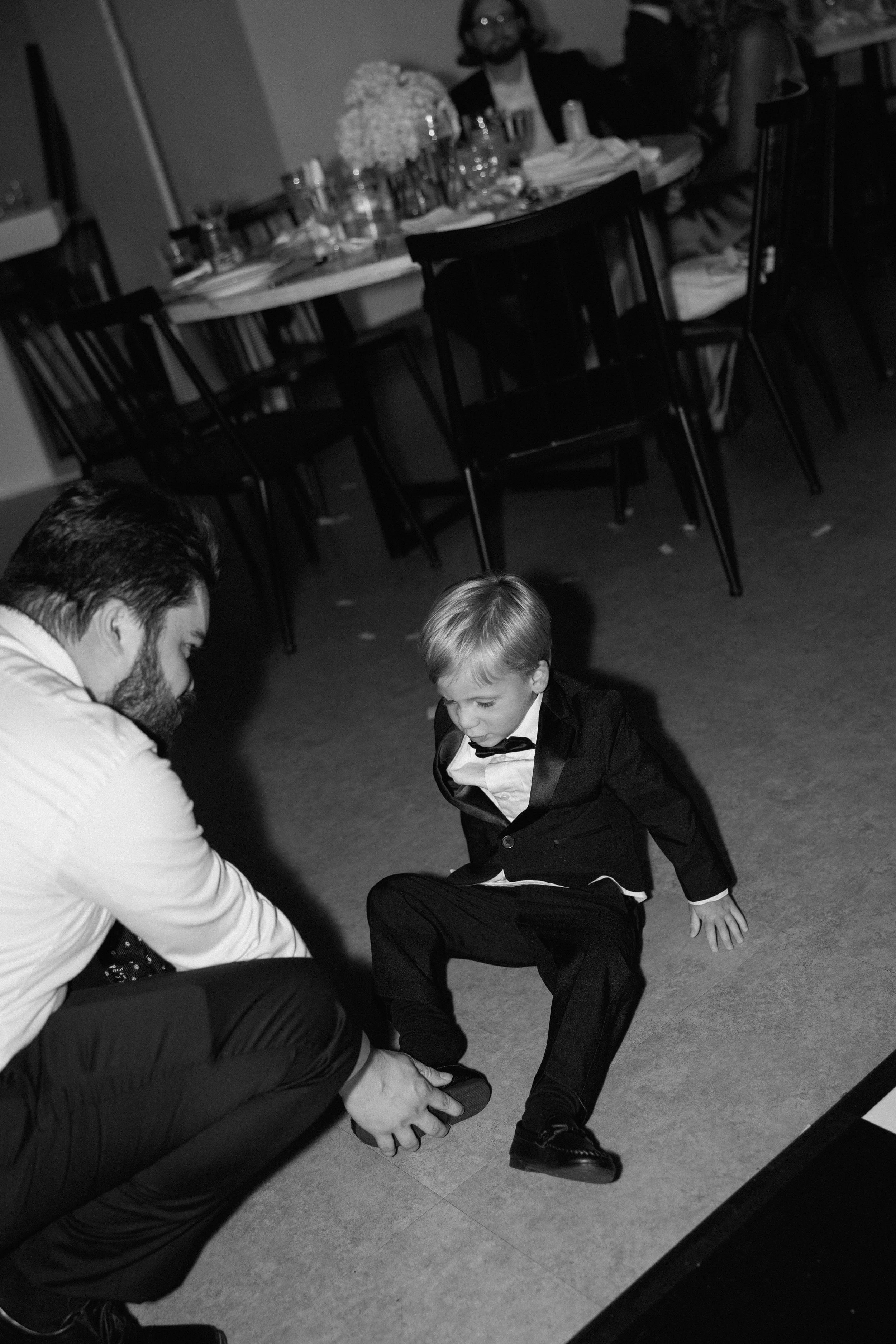 A young boy in a tuxedo sitting on the floor while an adult helps him put on his shoe at a formal event with tables set for dinner in the background.