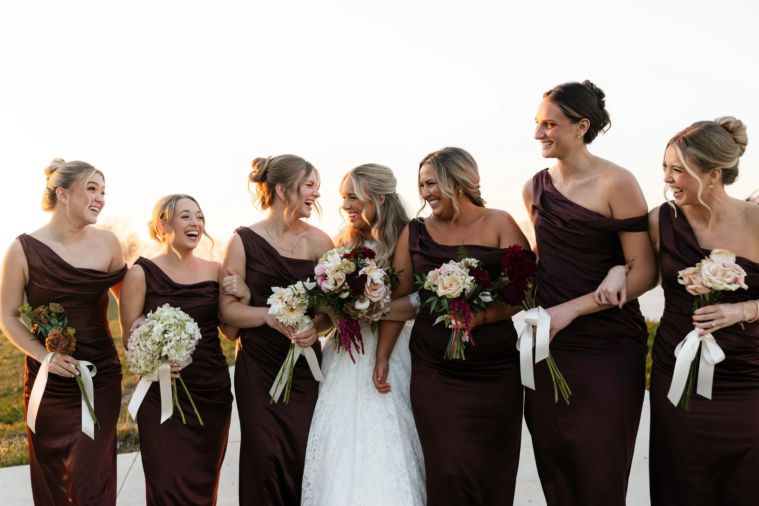 Bride and bridesmaids in matching dark purple dresses standing together outdoors during sunset, smiling and holding bouquets.