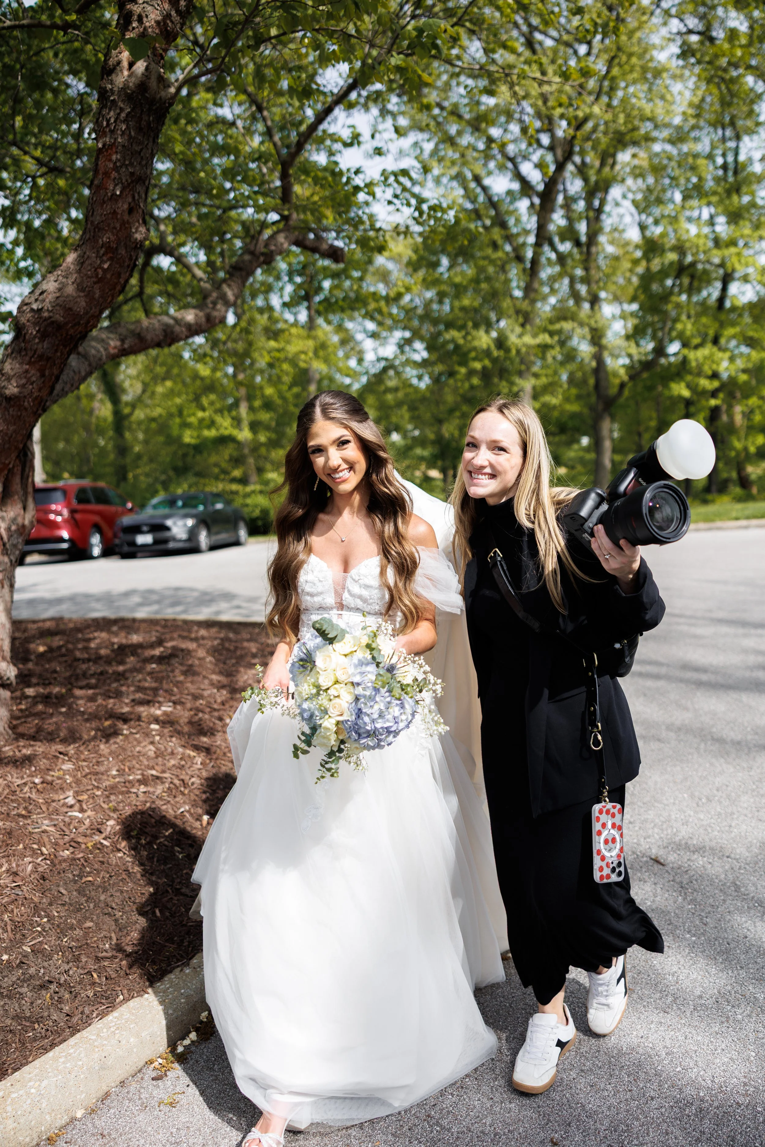 A bride in a white wedding dress holding a bouquet of white and blue flowers, smiling next to a photographer holding a camera, outdoors with green trees and parked cars in the background.