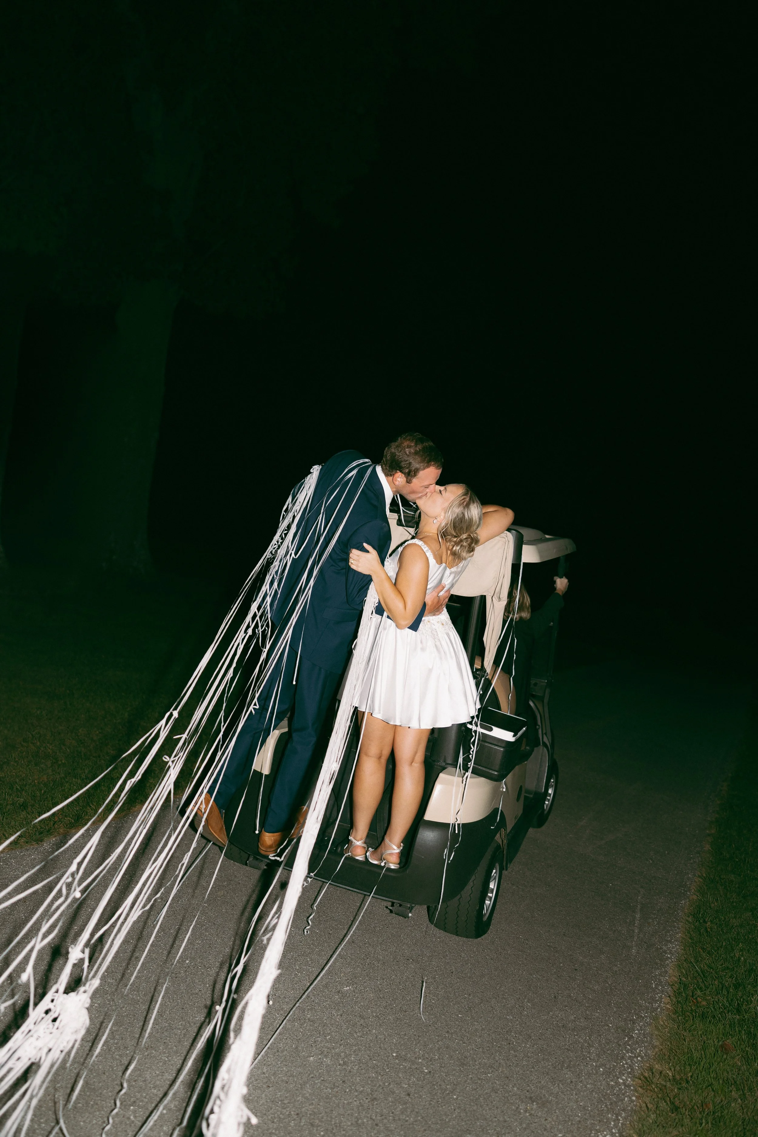A couple sharing a kiss on a golf cart at night, decorated with streamers and ribbons, with a person waving in the background.