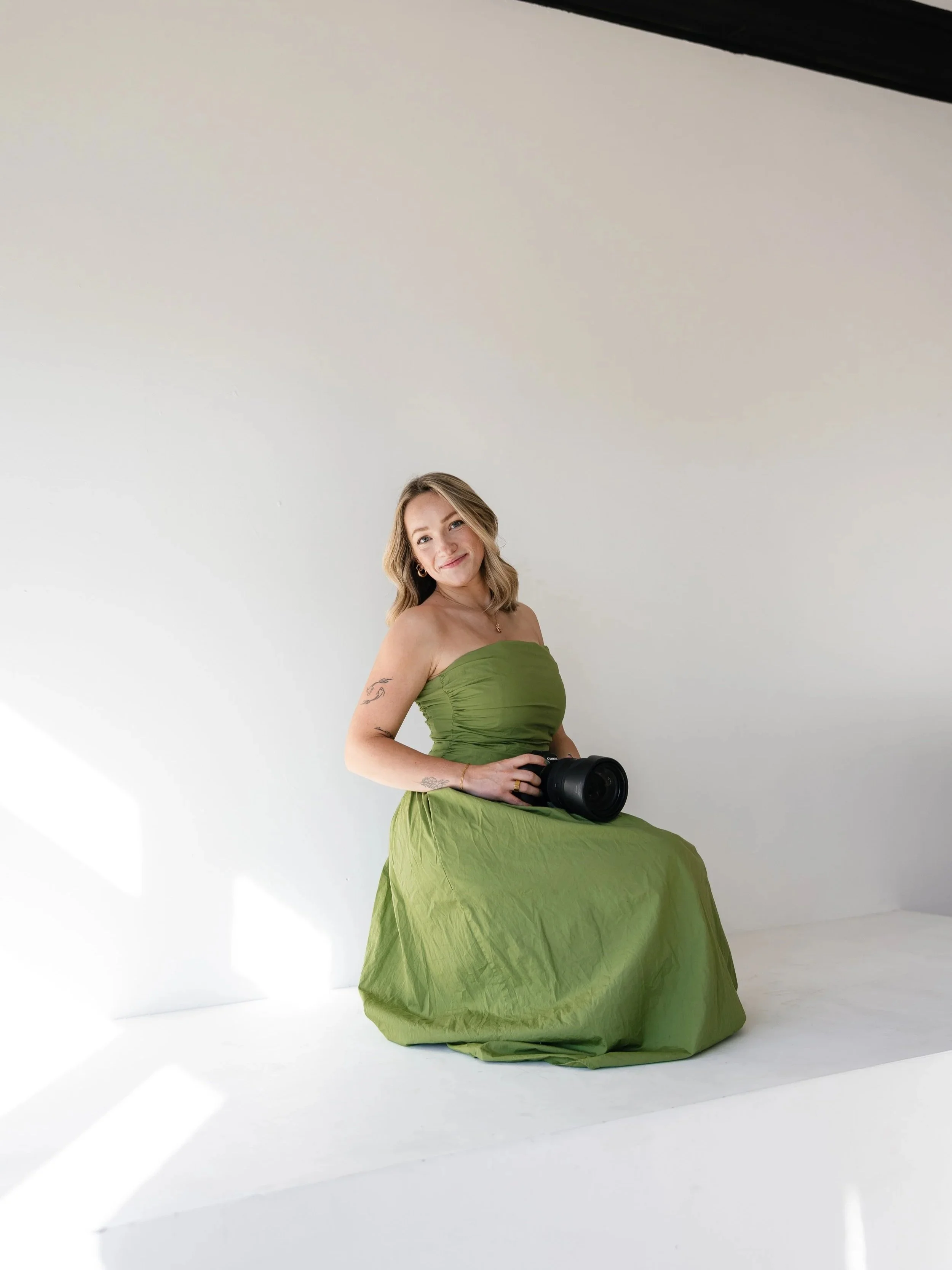 A woman in a green strapless dress sitting on the white floor, holding a camera, smiling at the camera, against a plain white background.