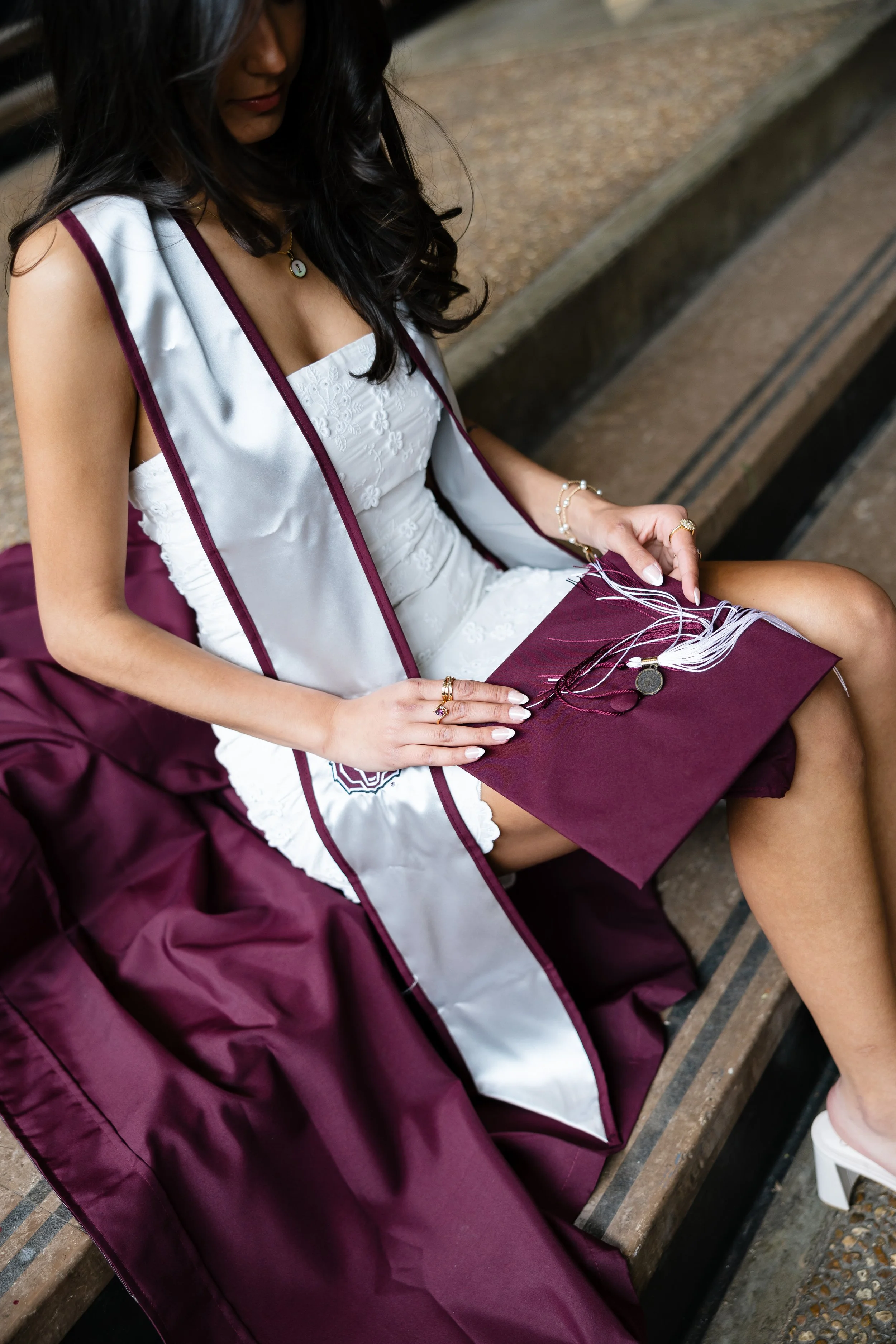 A woman in a white dress and maroon graduation gown holding a maroon diploma cover and tassel, sitting on a wooden bench.