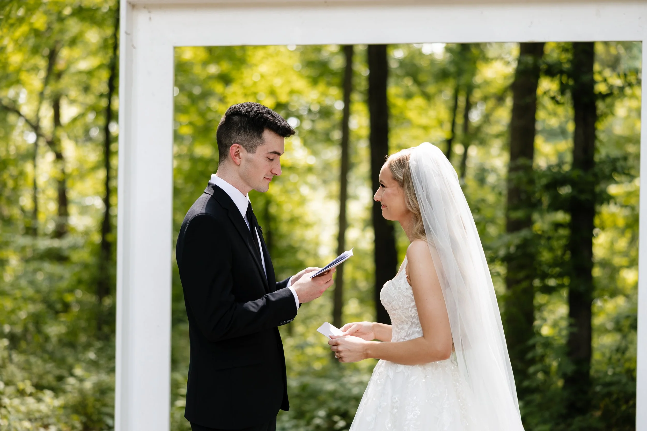 A bride and groom facing each other during their wedding ceremony outdoors among trees, with the groom reading vows from a small book.