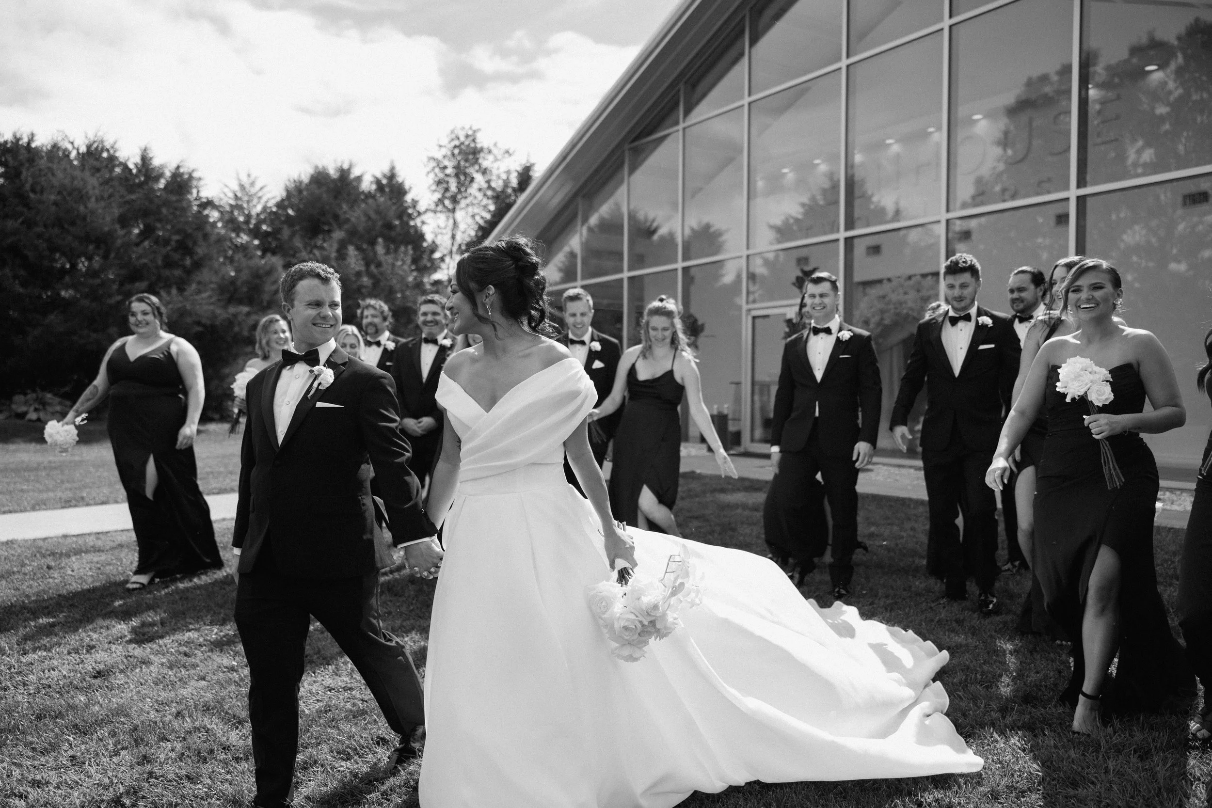 A bride and groom stand outside a white barn decorated for a wedding, with the bride in a white dress and the groom in a black suit, holding hands and looking at each other.