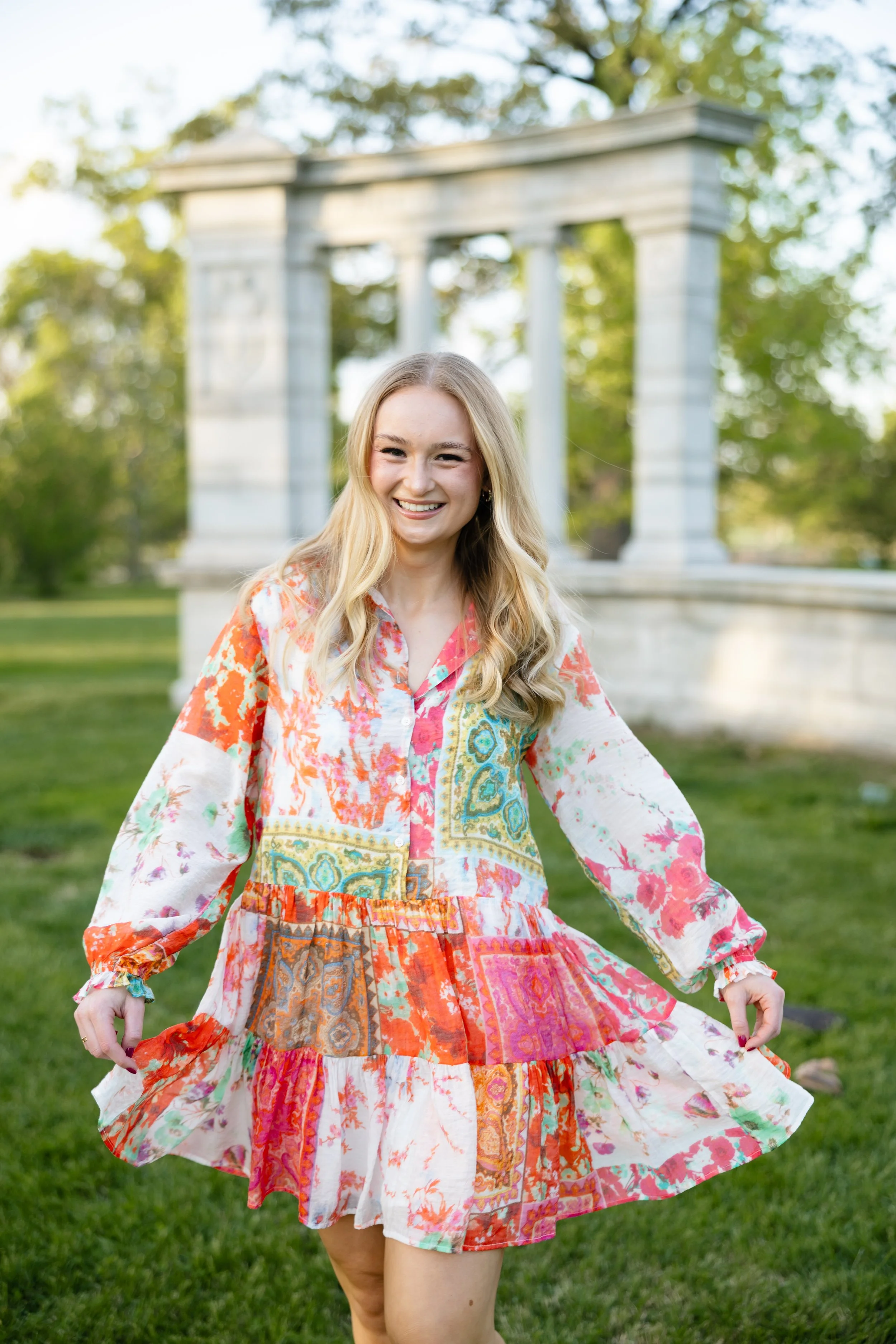 A young woman with blonde hair in a colorful, patterned dress smiling outdoors in front of a stone structure with trees in the background.