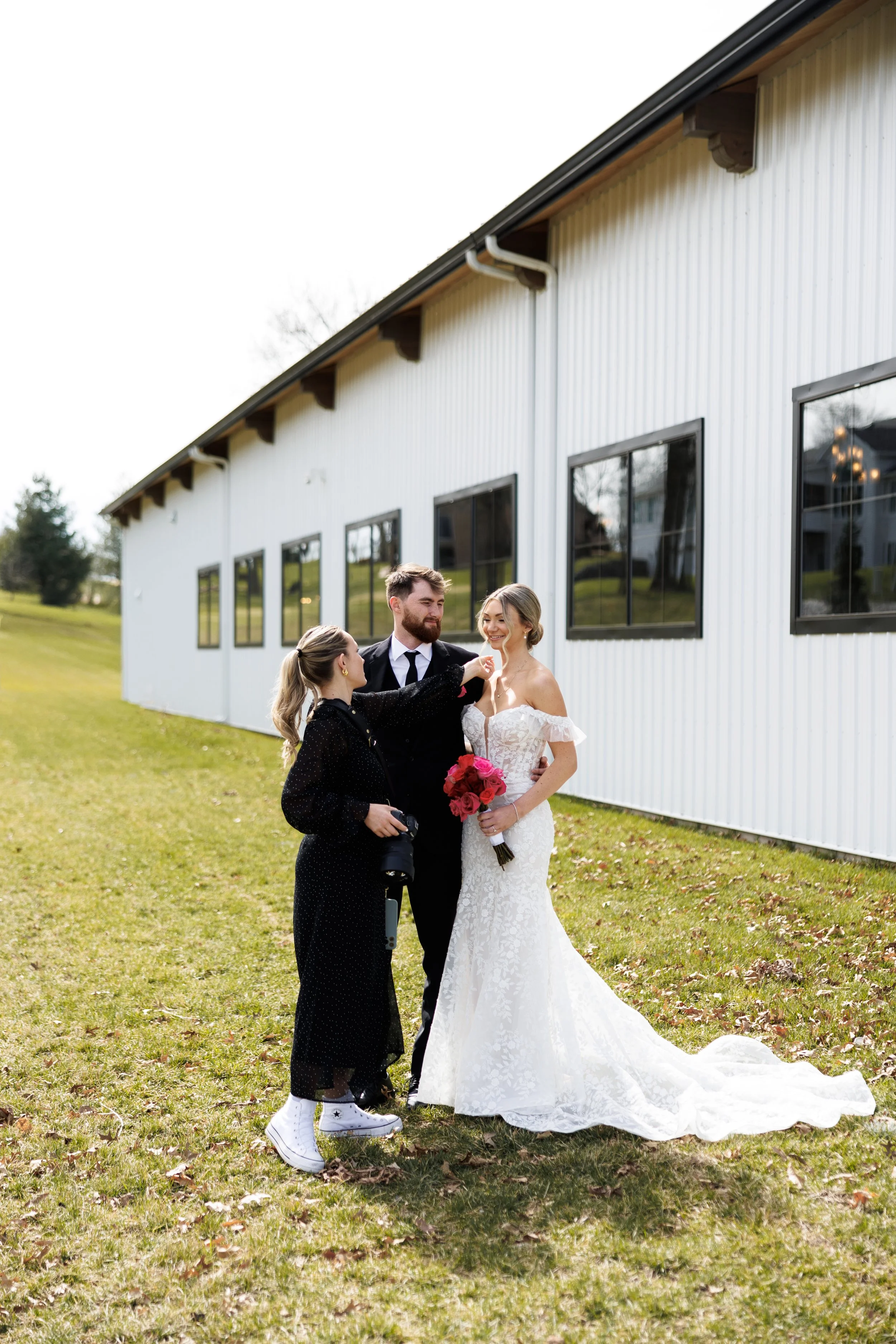 A bride and groom on their wedding day outside next to a white building, with a woman holding a camera, smiling and pointing at the bride.