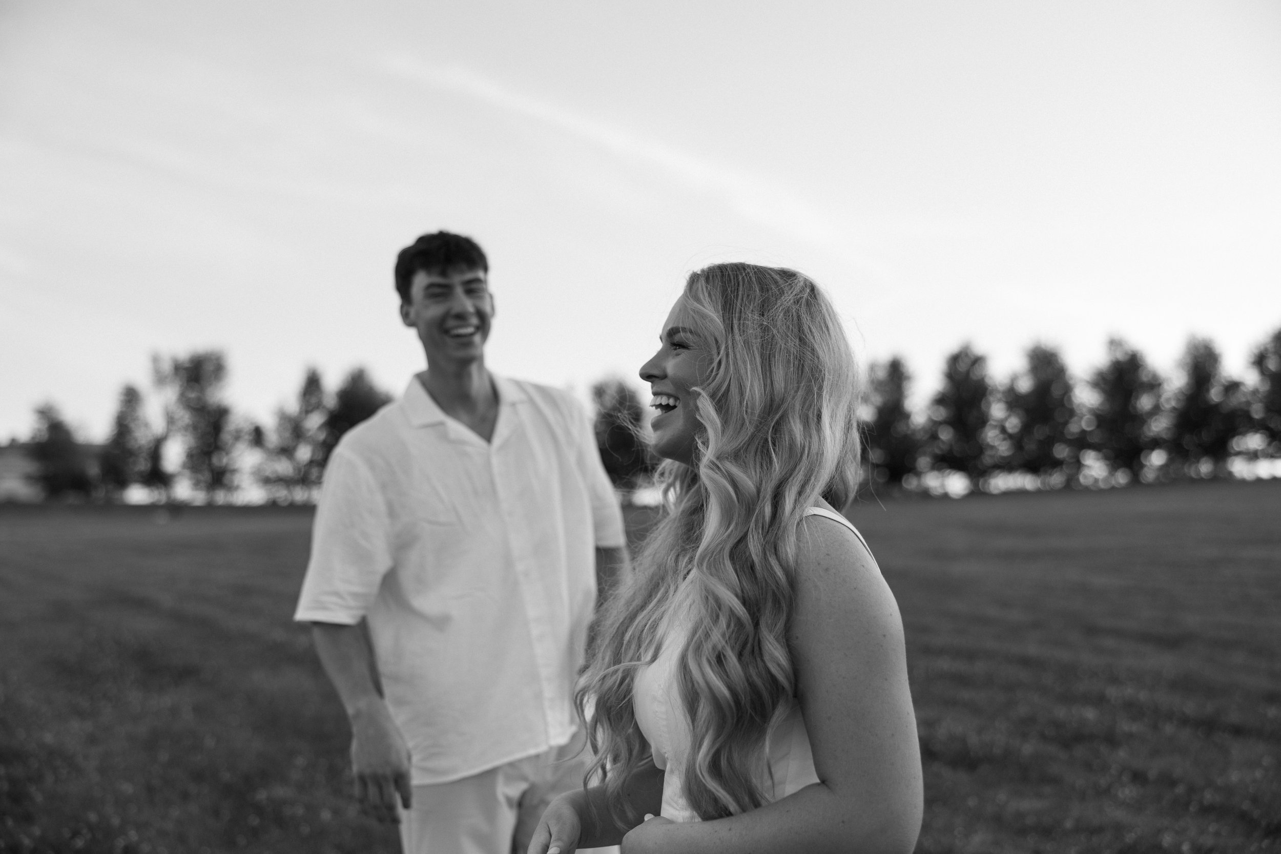 Black and white photo of a woman with long wavy hair laughing and a man in the background smiling in an open field with trees in the distance.
