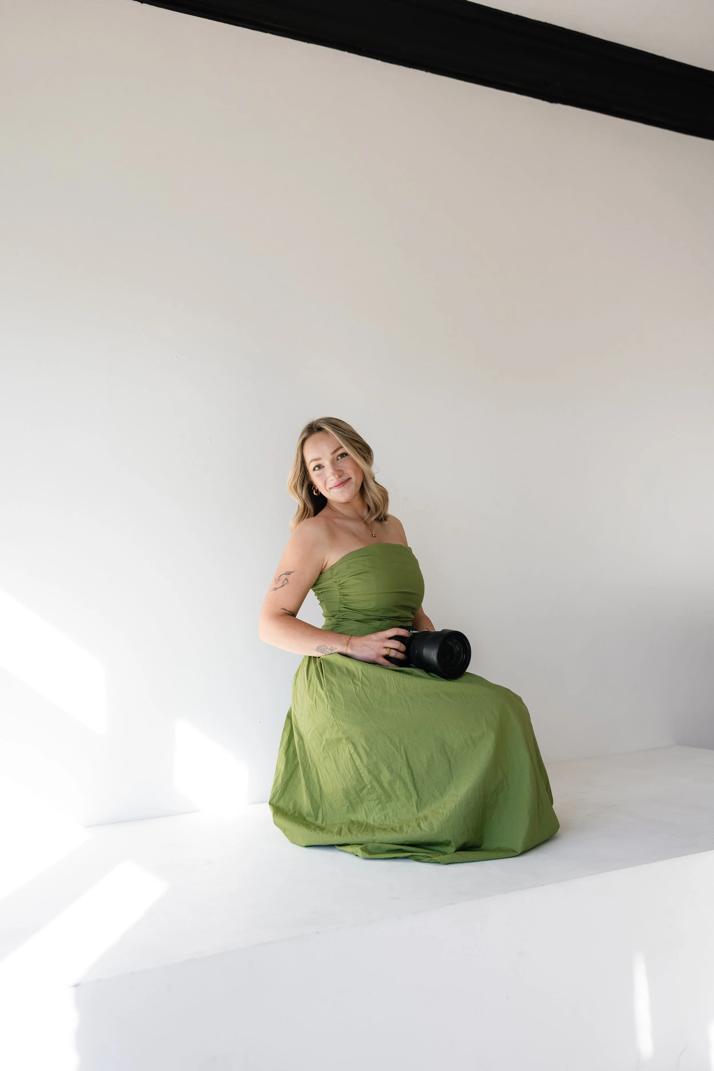A woman in a green strapless dress sitting on a white platform in a room with white walls, holding a camera, smiling at the camera.