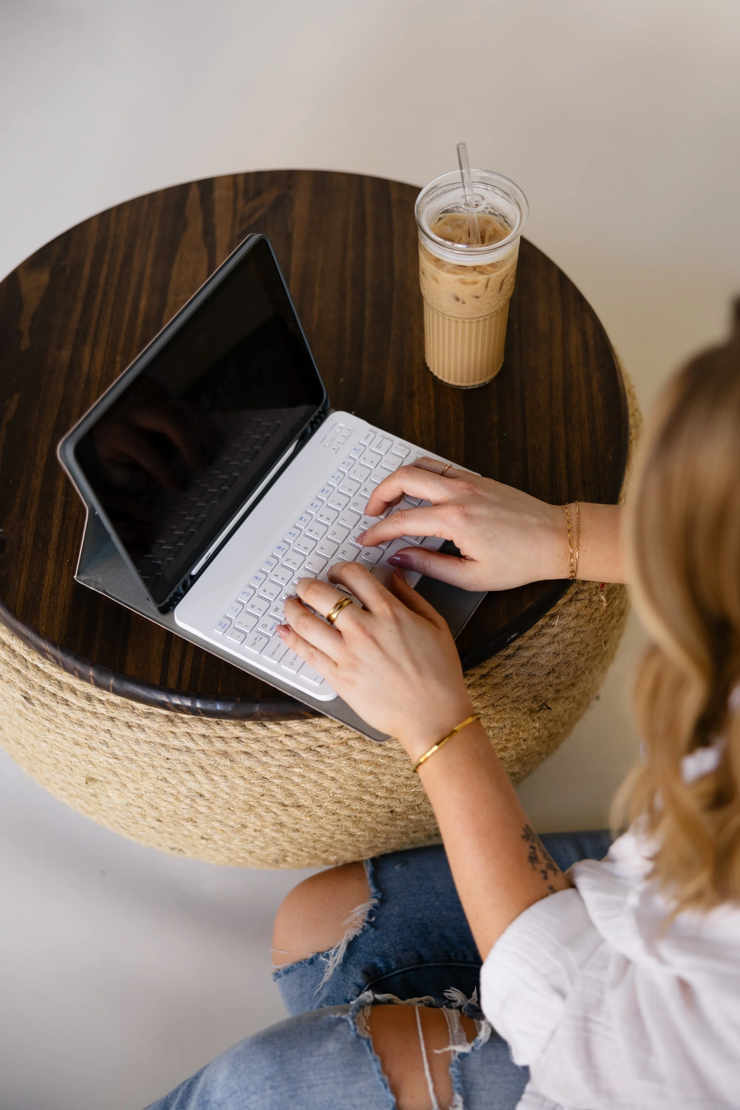 Person using a white keyboard laptop on a round wooden table, with a tall iced coffee drink nearby.