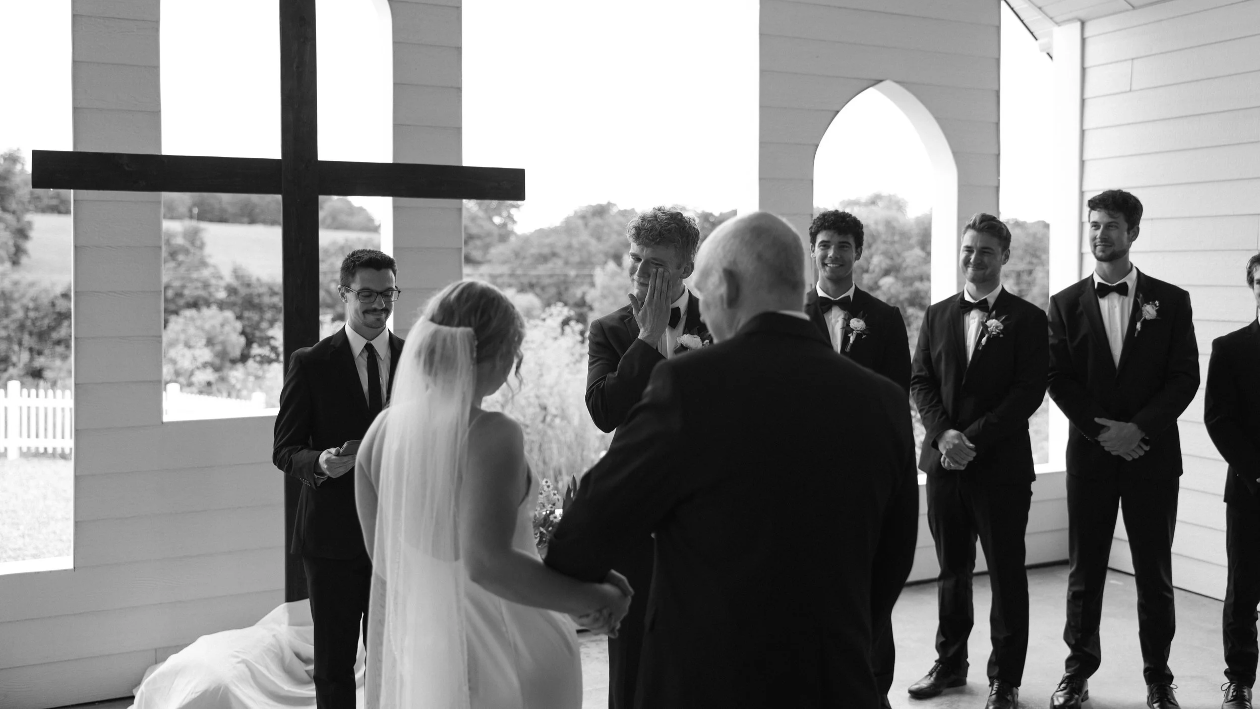 Black and white photo of a woman with long wavy hair laughing and a man in the background smiling in an open field with trees in the distance.