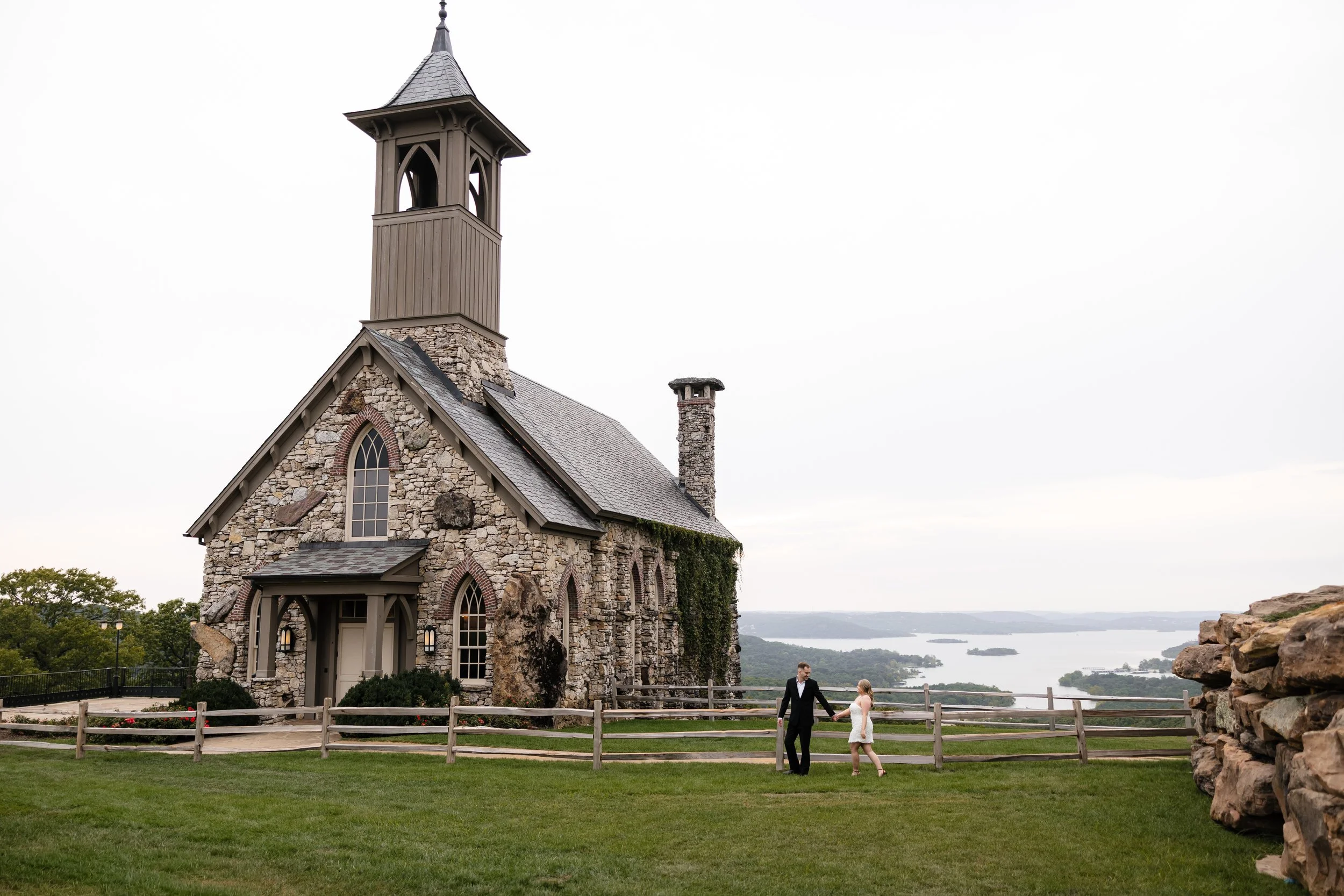 A man and woman in wedding attire holding hands outside a stone church with a bell tower, overlooking a lake and landscape.
