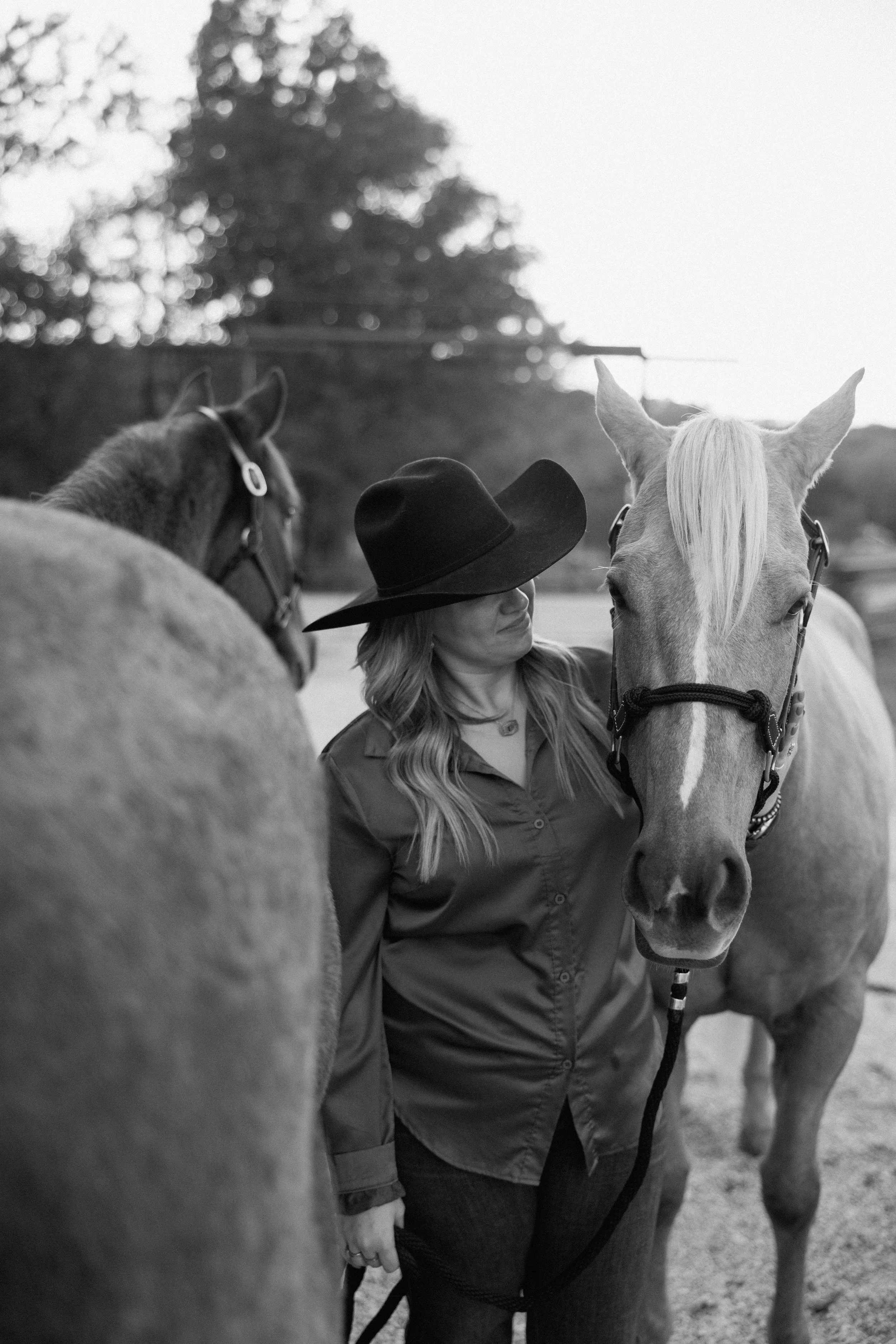 A woman in a cowboy hat and button-up shirt standing between two horses, one facing the camera and the other with its back turned, outdoors.