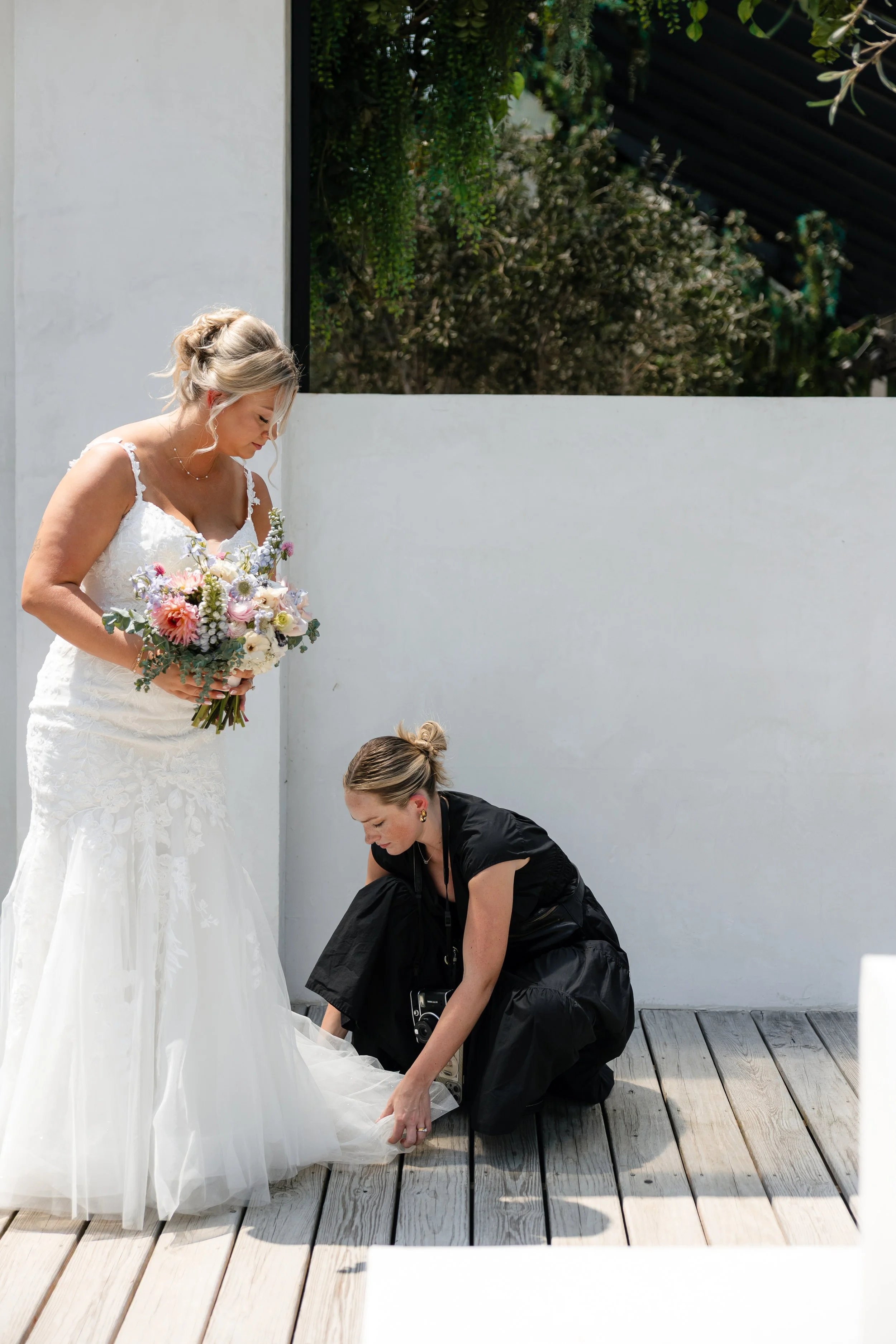 A bride holding a bouquet looks down at a woman adjusting her dress on a wooden deck outside.