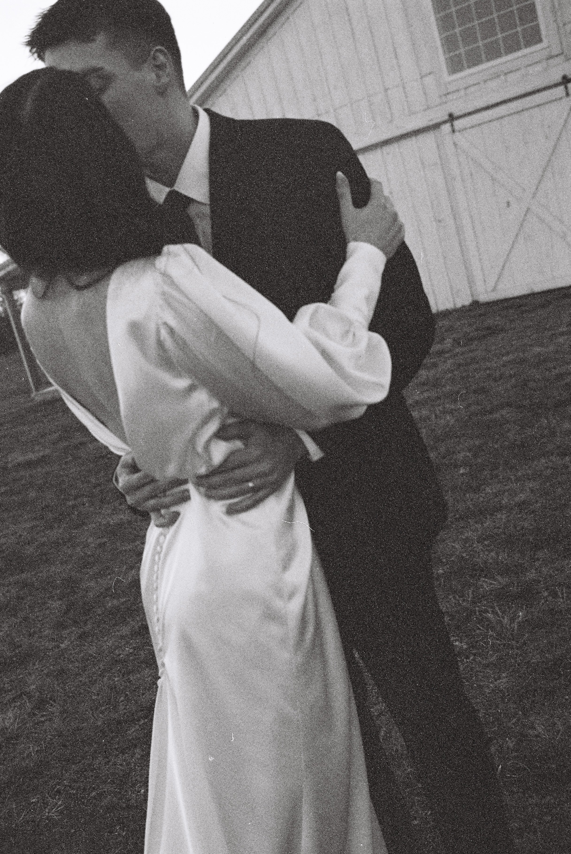 A black-and-white photo of a couple kissing outdoors, with a barn on the background. The man is dressed in a suit, and the woman is wearing a dress.