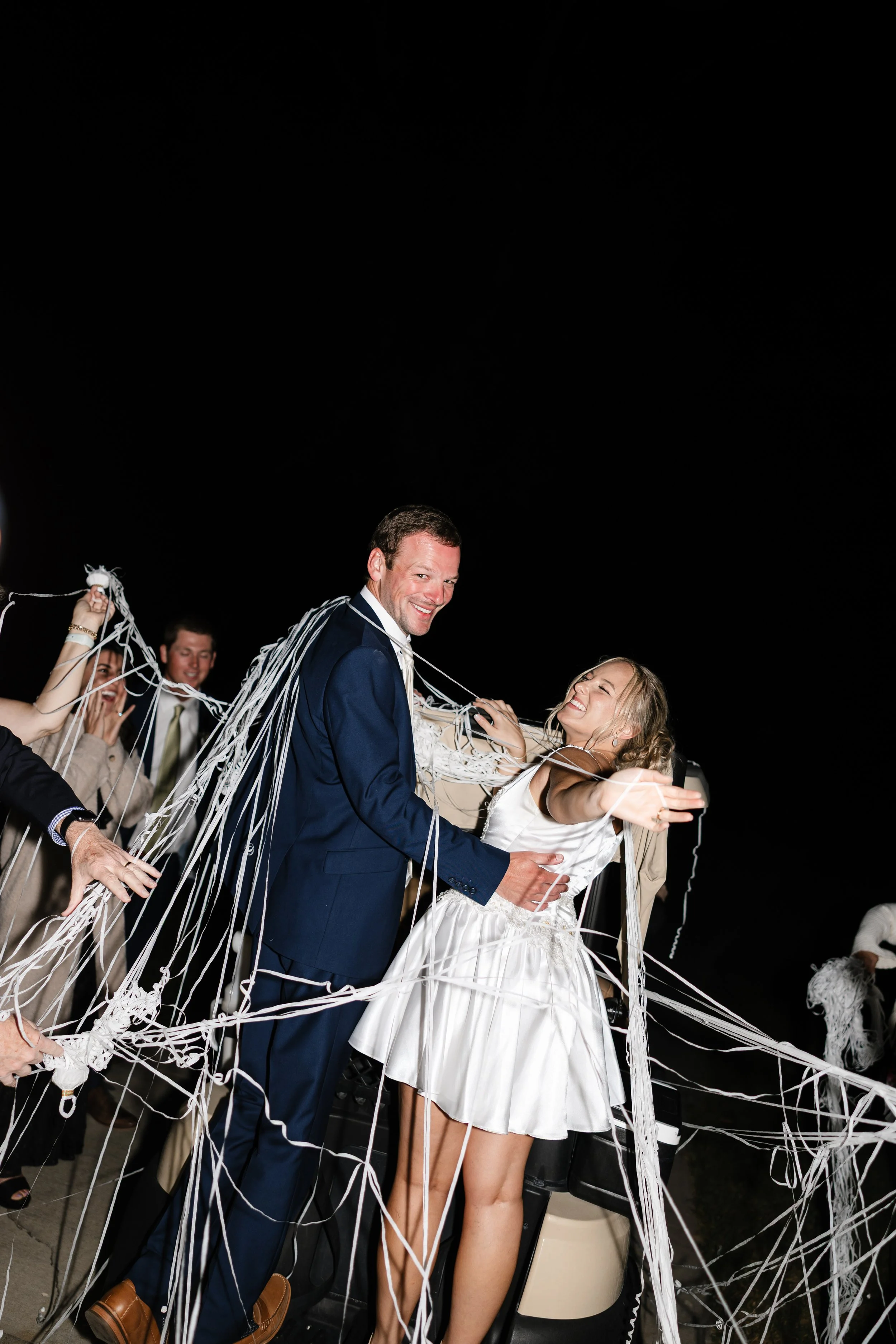 A bride and groom smiling and hugging during their wedding celebration surrounded by friends pulling white streamers in the night.