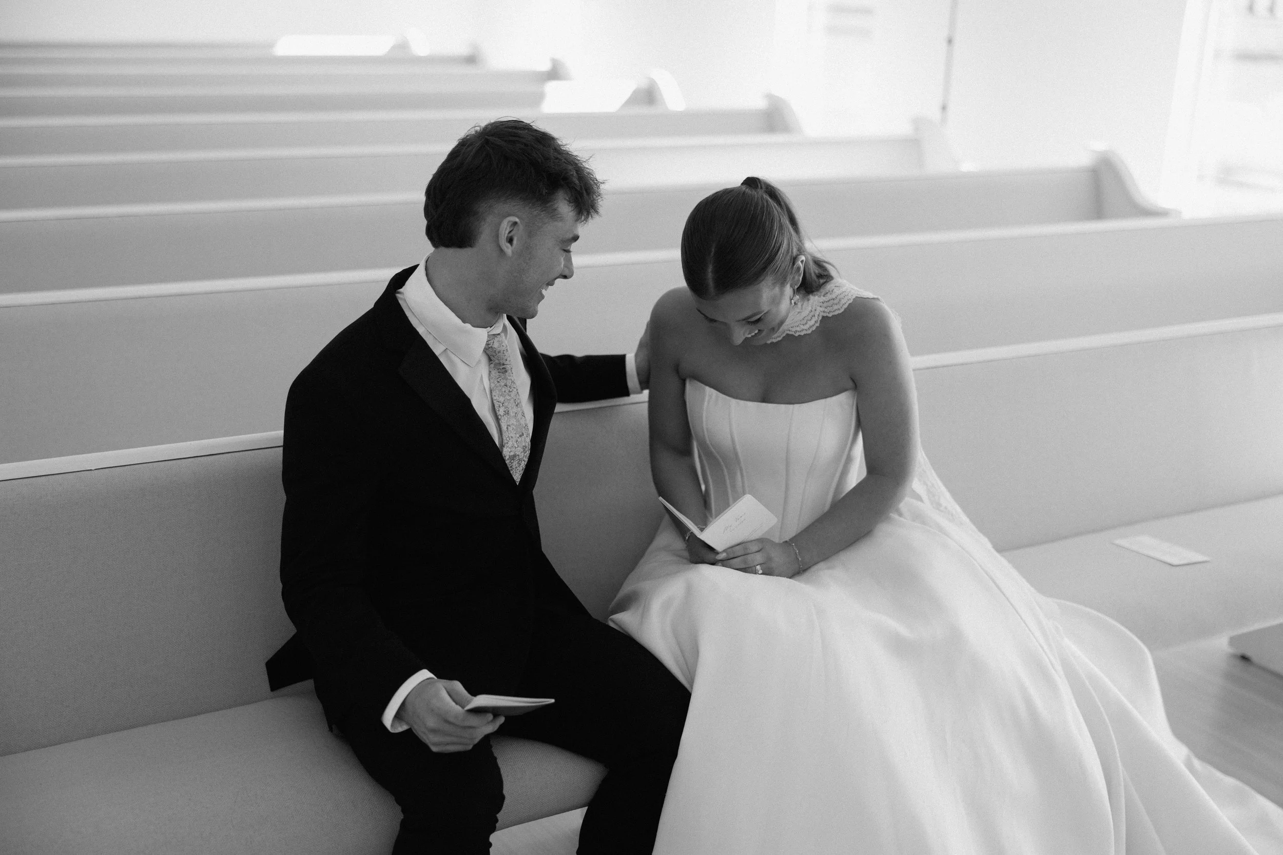 A bride and groom sitting on a church bench, looking at a booklet together, smiling and laughing.